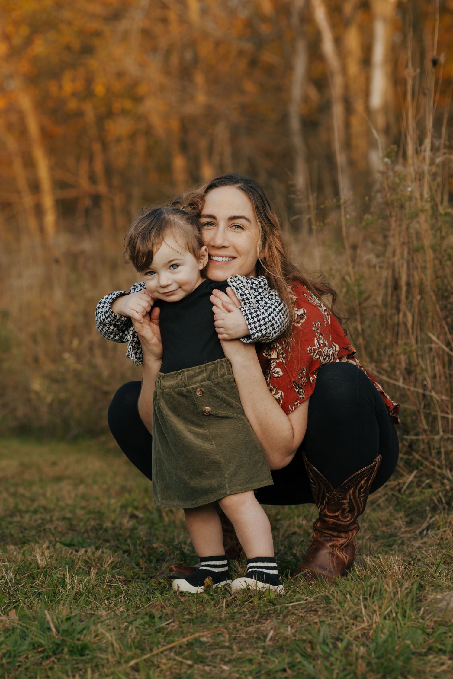 Smiling mother crouching to hug young toddler girl in green skirt during fall outdoor portrait.