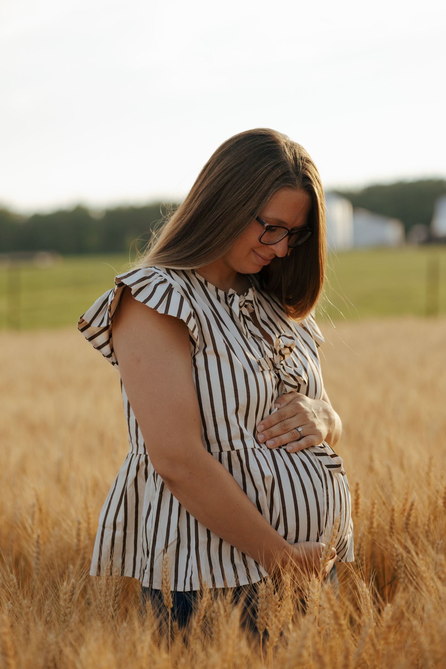 Pregnant woman in striped dress cradling baby bump in golden wheat field at dusk.