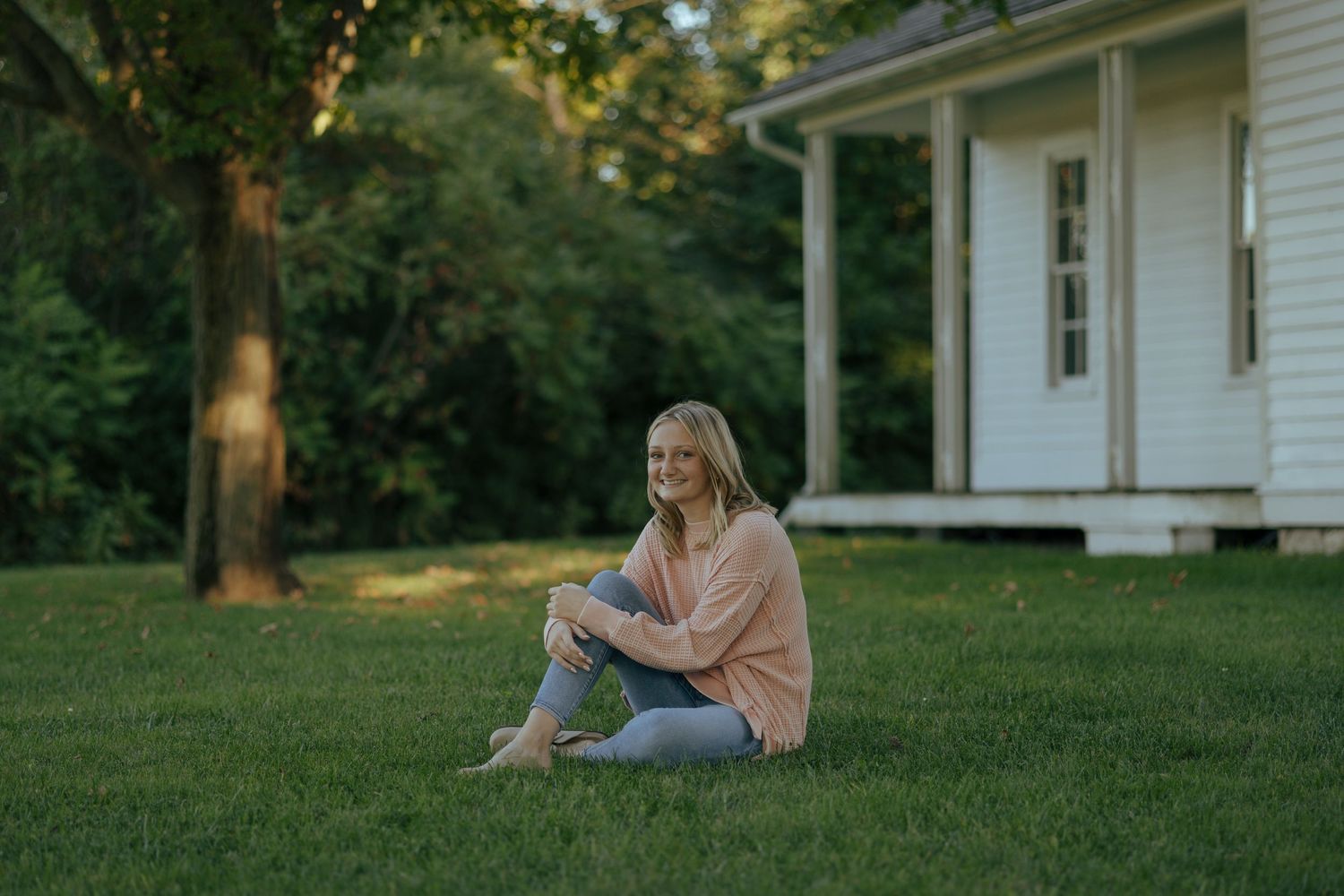 Smiling blonde teen in pink sweater and jeans sitting barefoot on grass near white farmhouse.