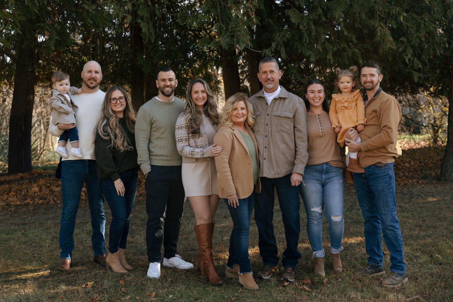 Extended family group portrait smiling together outdoors among pine trees in autumn.