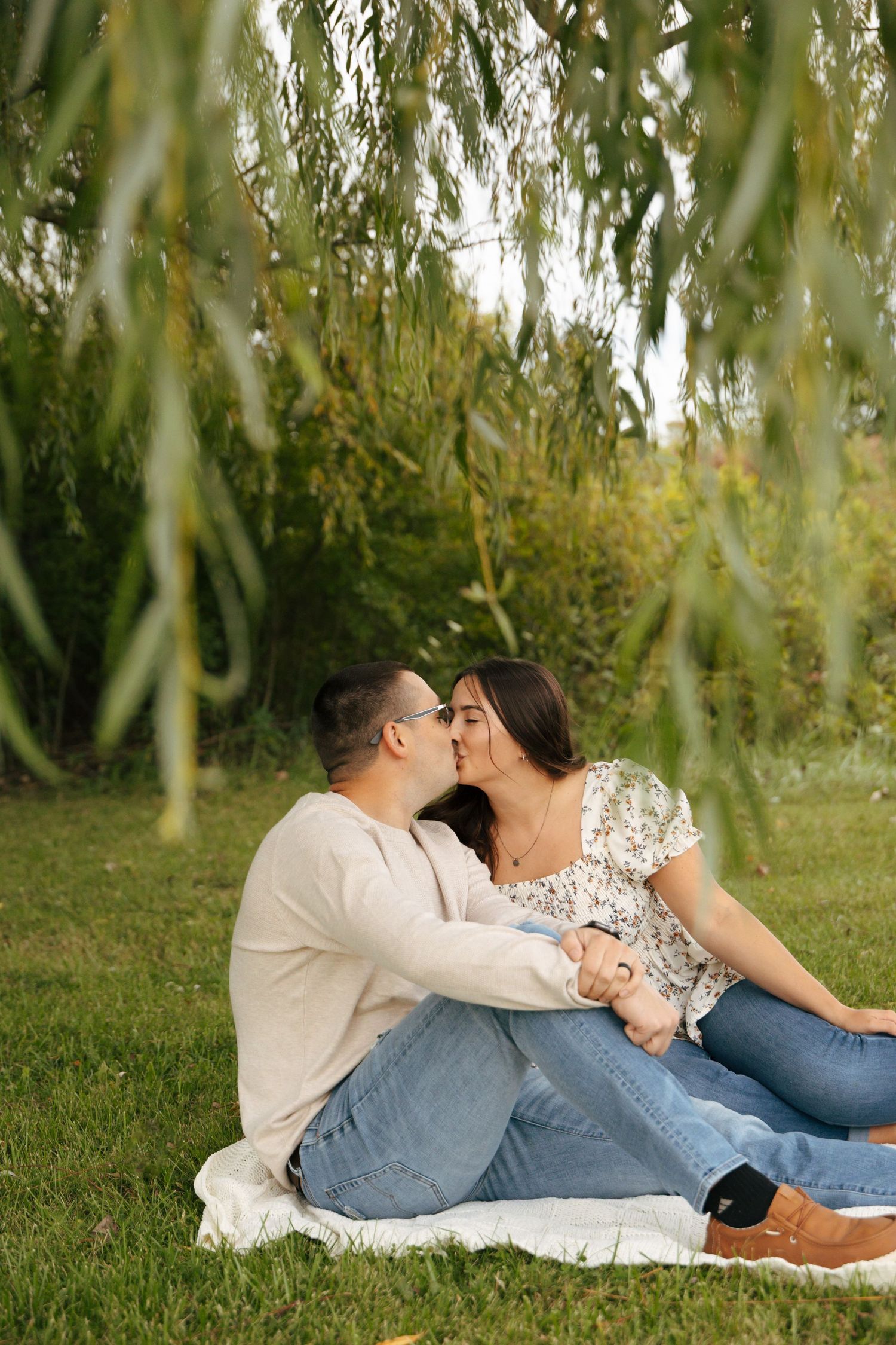 Couple kissing while sitting on a blanket in a lush green park under a willow tree.