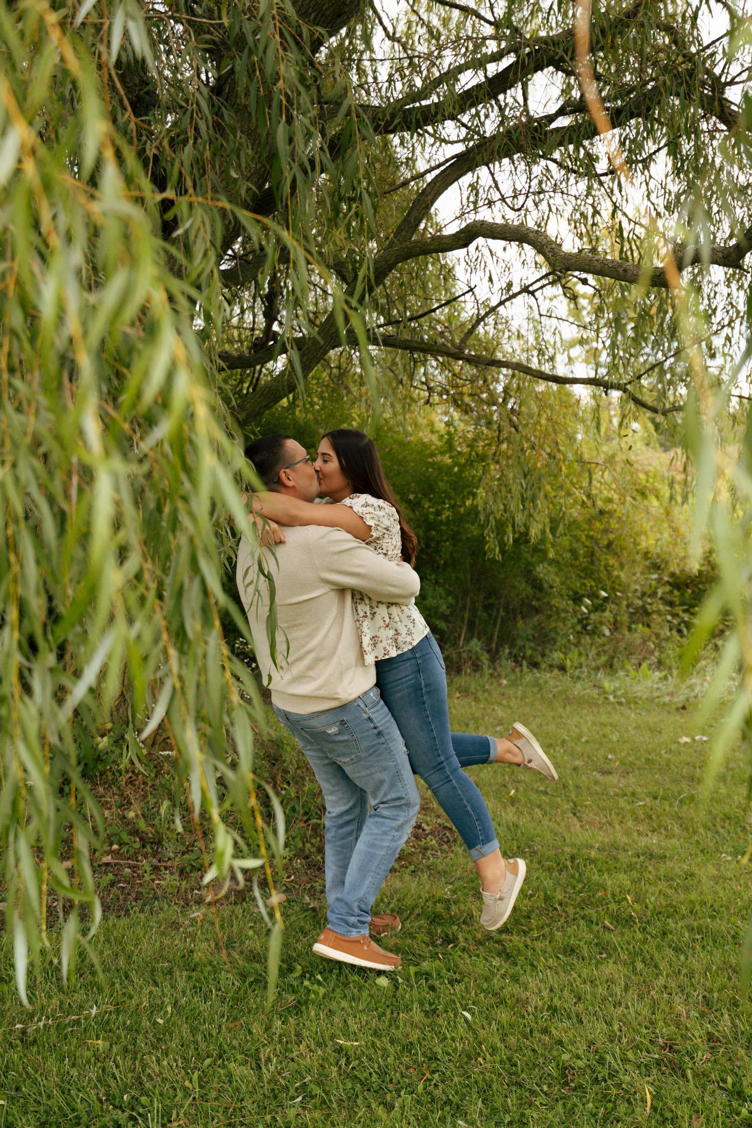 Couple kissing passionately while embracing under a weeping willow tree in a lush park.