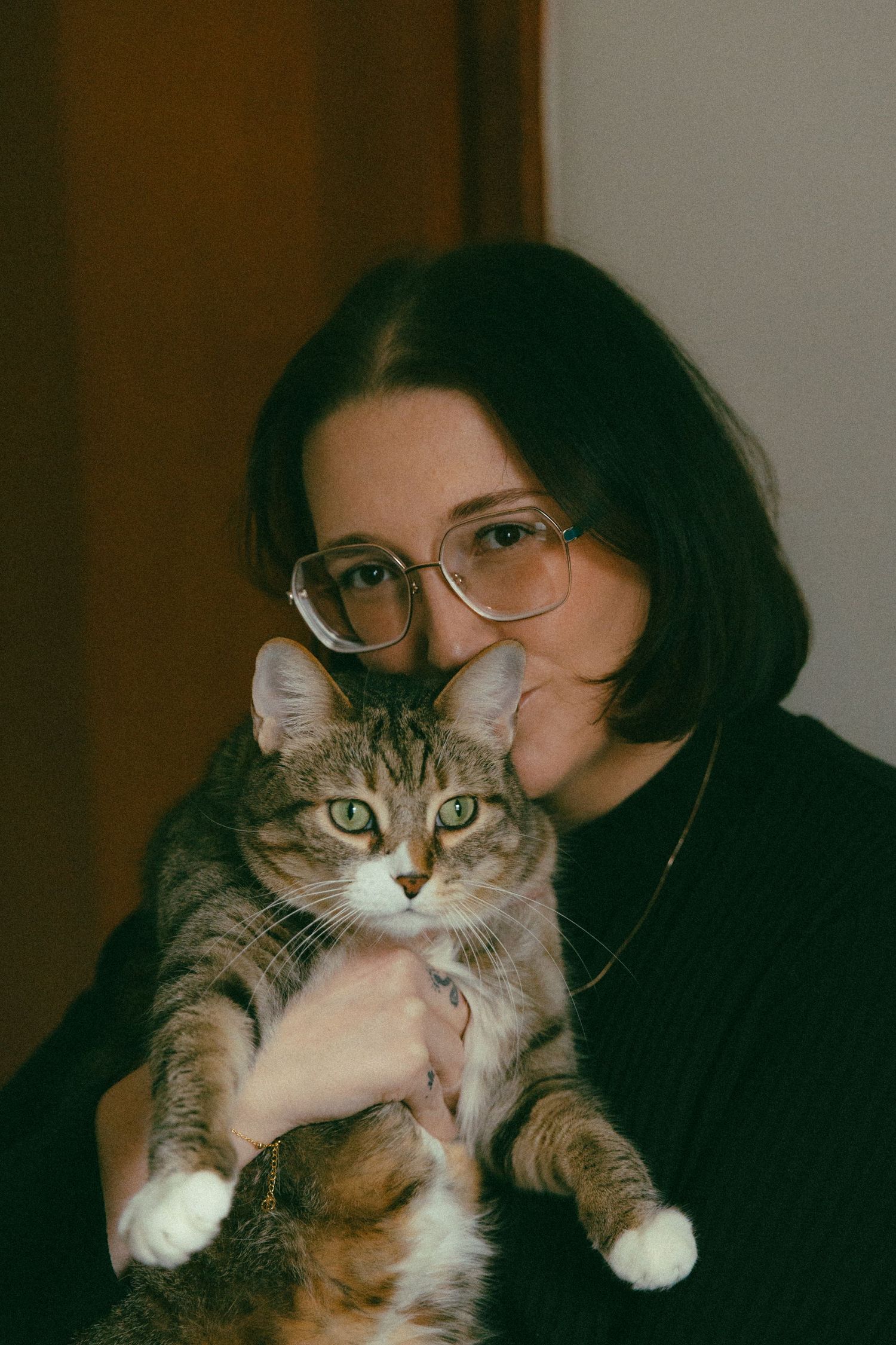 Young woman with glasses in black sweater holding a tabby cat with green eyes indoors.