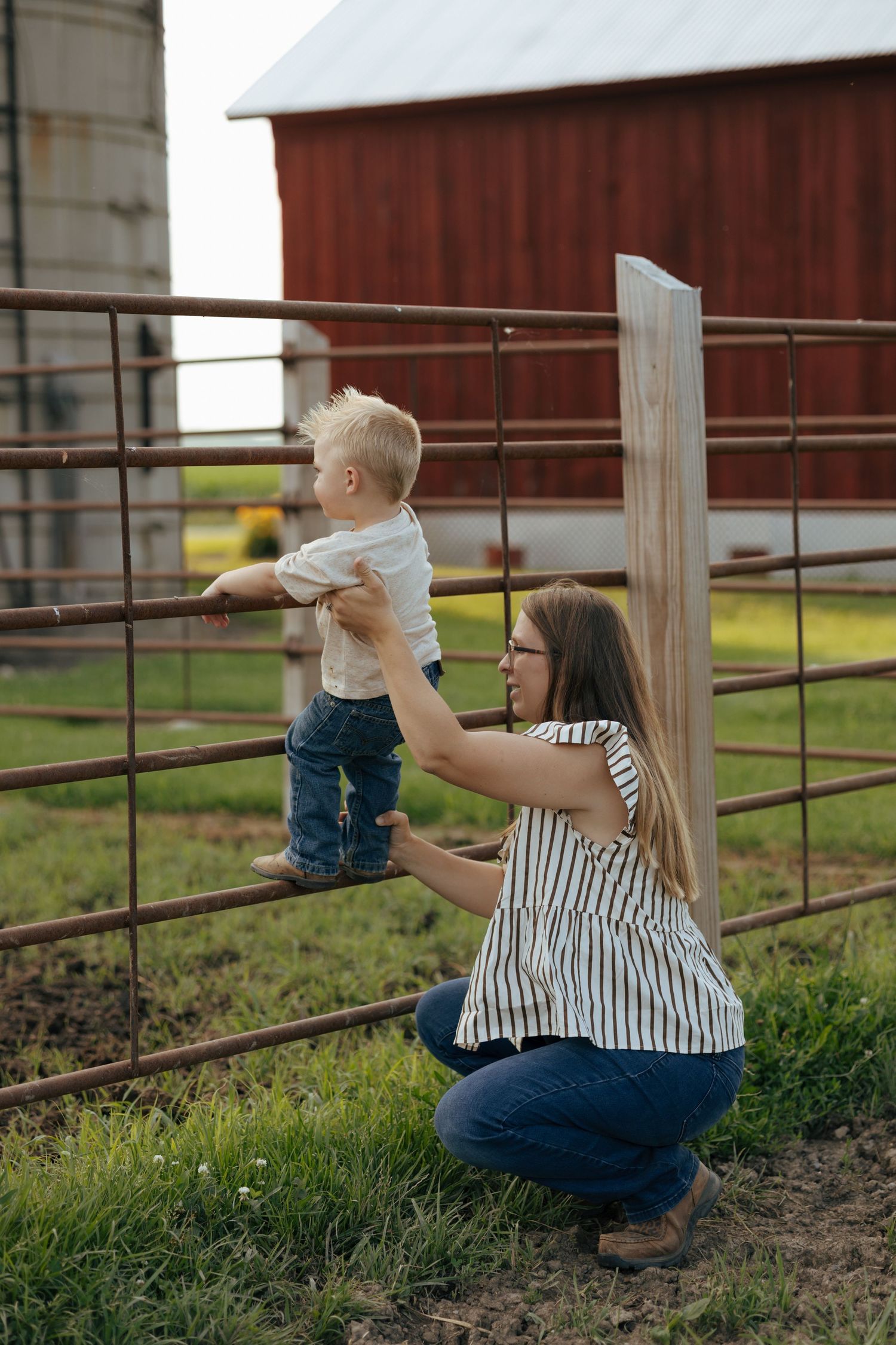 Mother helping young toddler climb a rustic farm gate near a red barn and silo.