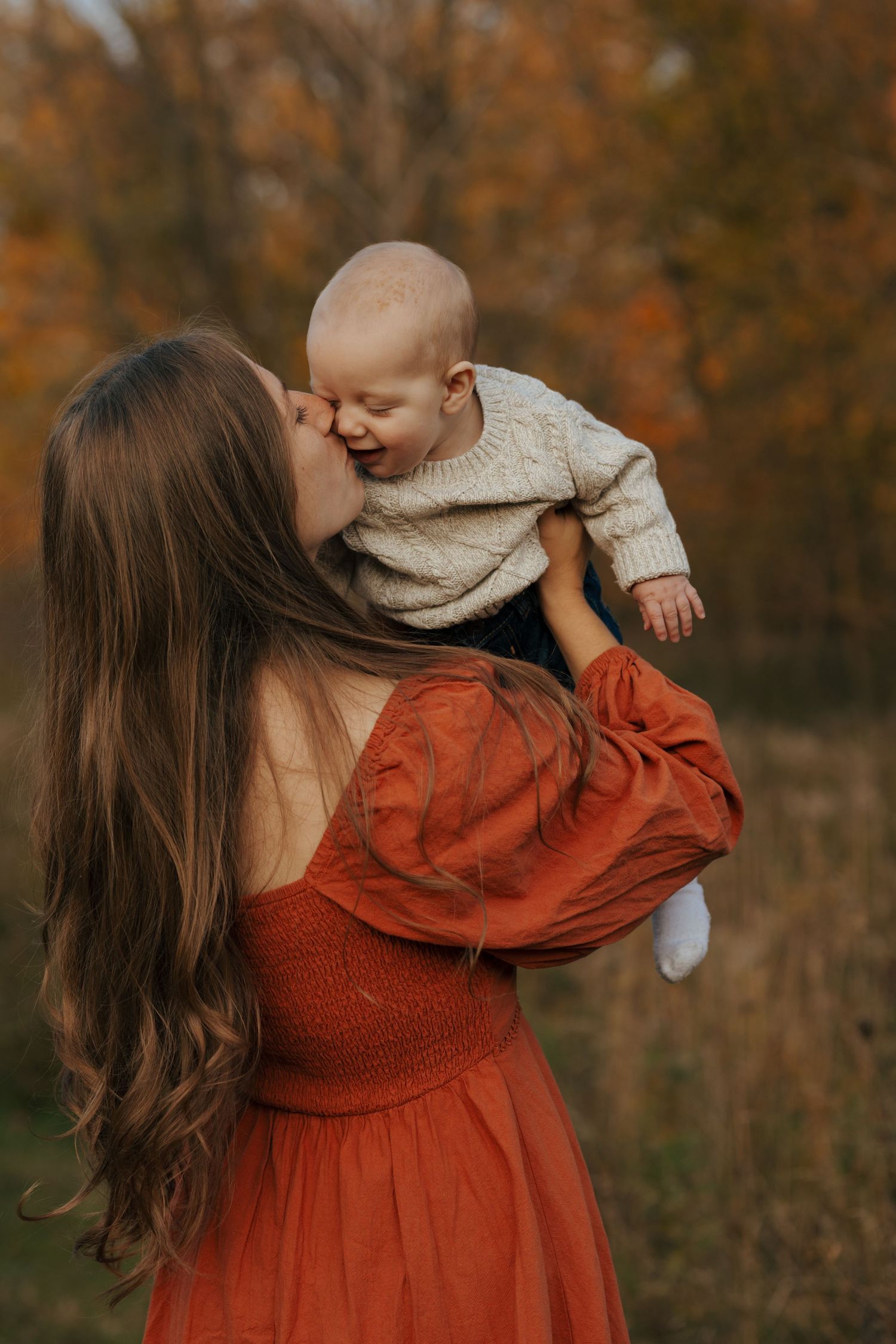 Mother in rust red dress kissing laughing baby held up in autumn outdoor portrait session.
