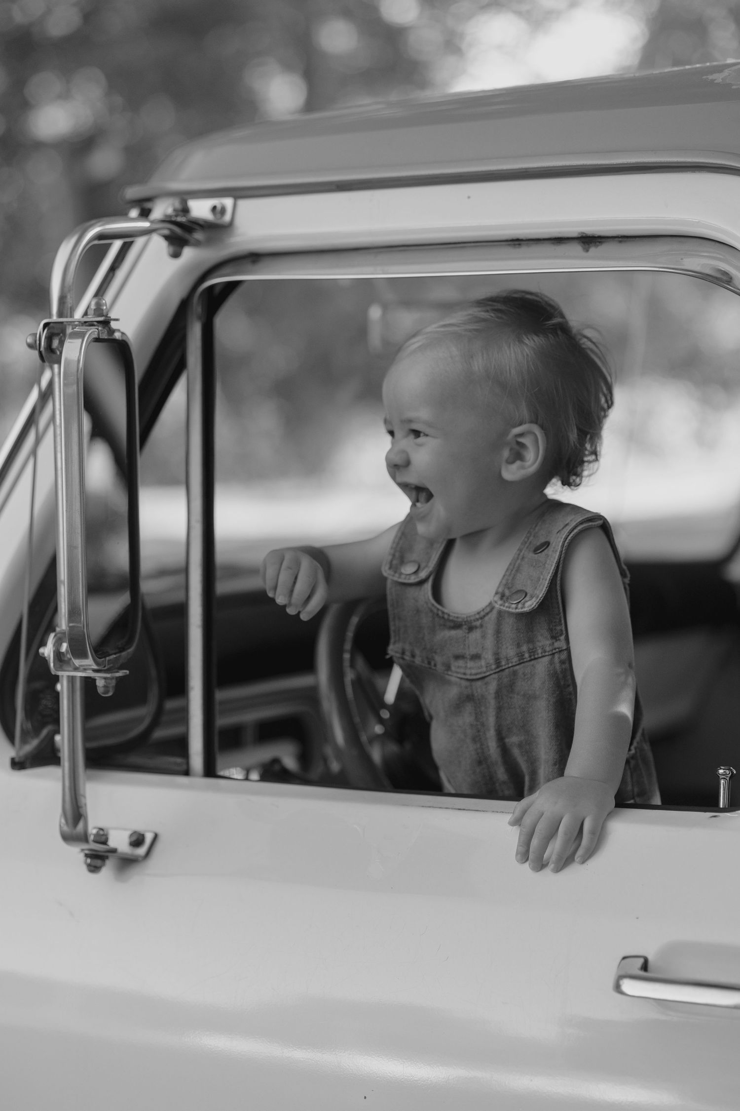 Laughing baby in denim overalls leaning out window of vintage truck, black and white.