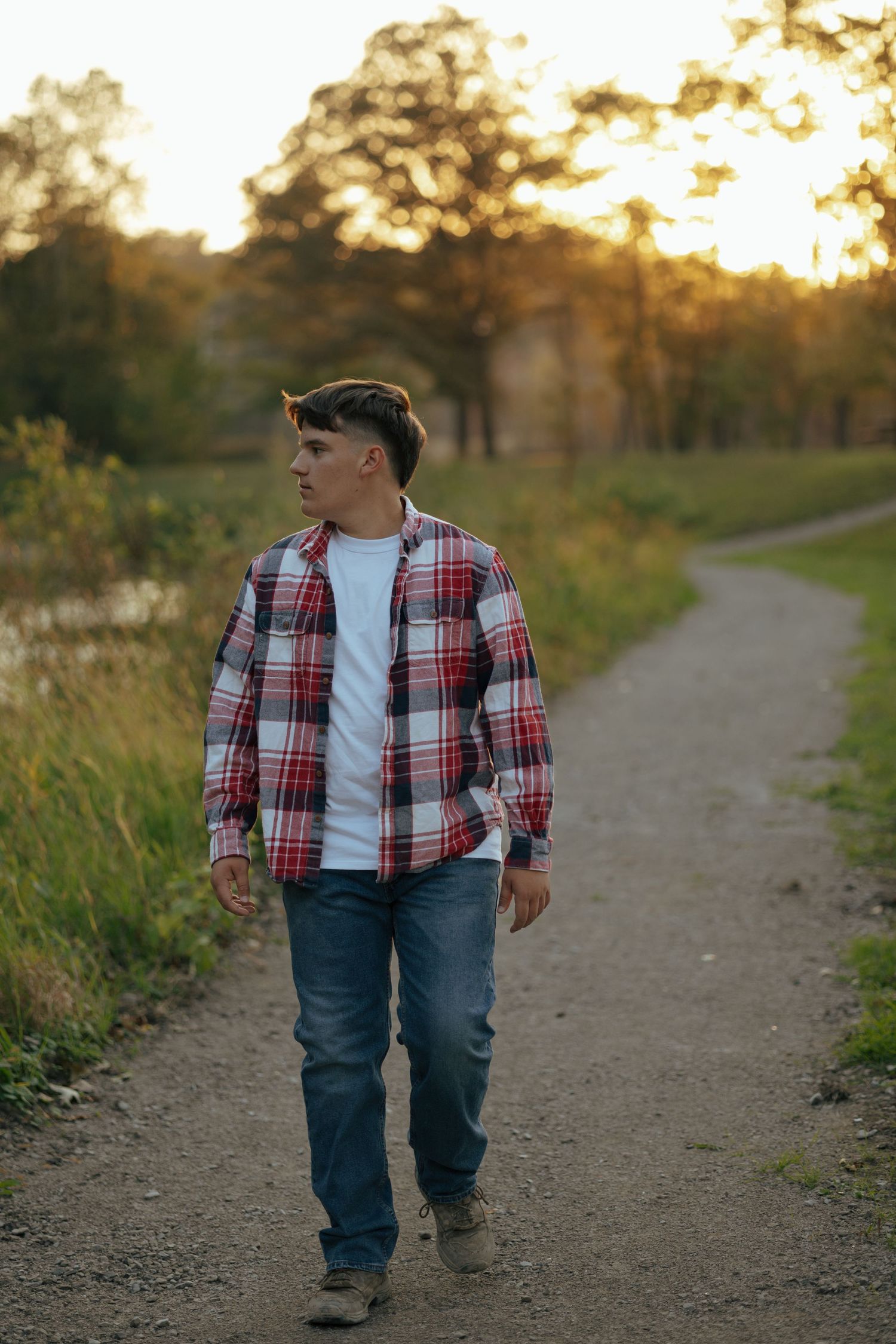 Teen boy in red plaid flannel shirt and jeans walks gravel park path at golden sunset.