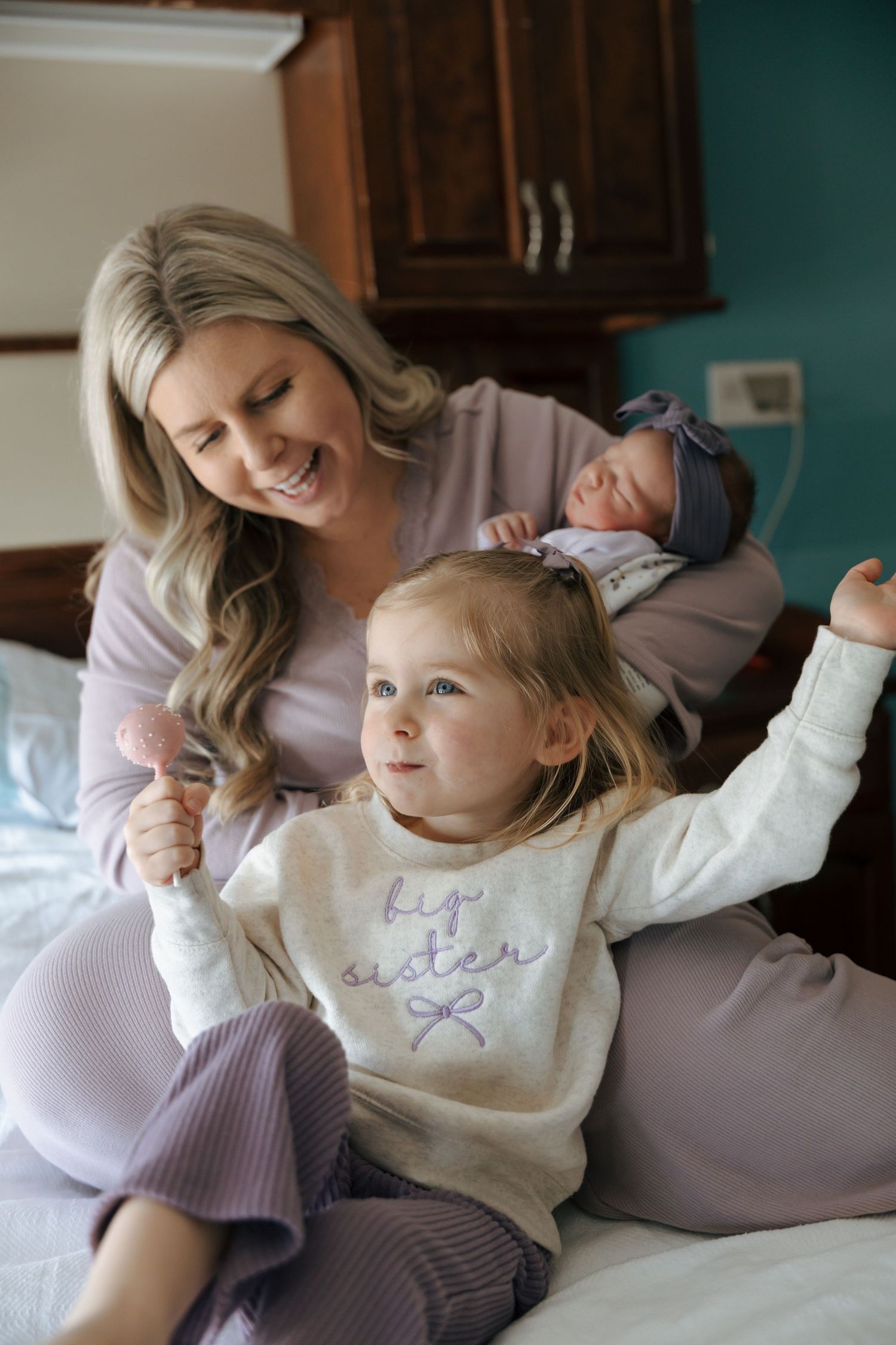 Smiling mother holding newborn while toddler in big sister sweatshirt plays on hospital bed.