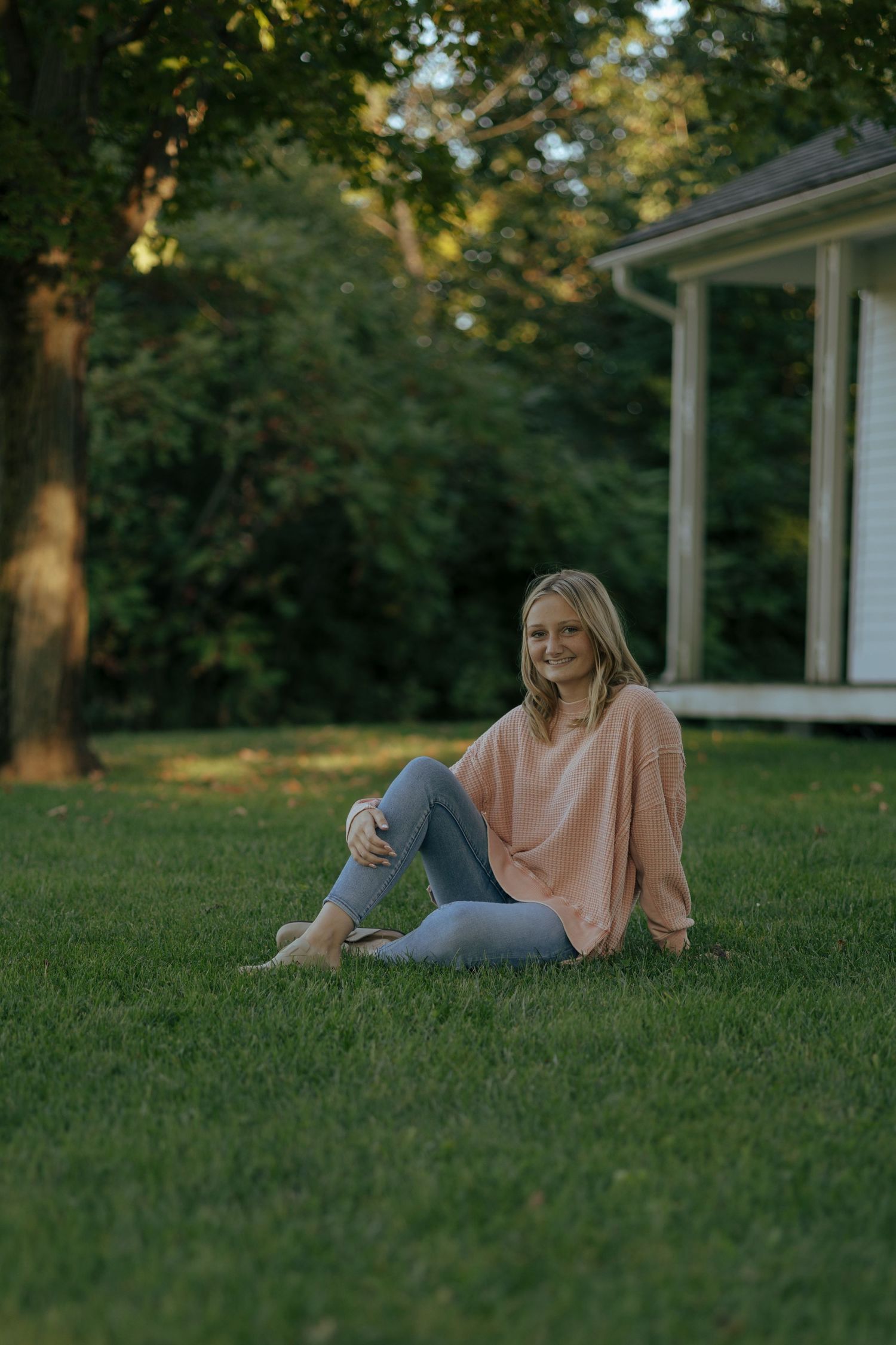 Smiling blonde teenage girl sitting on green grass lawn wearing pink sweater and jeans.