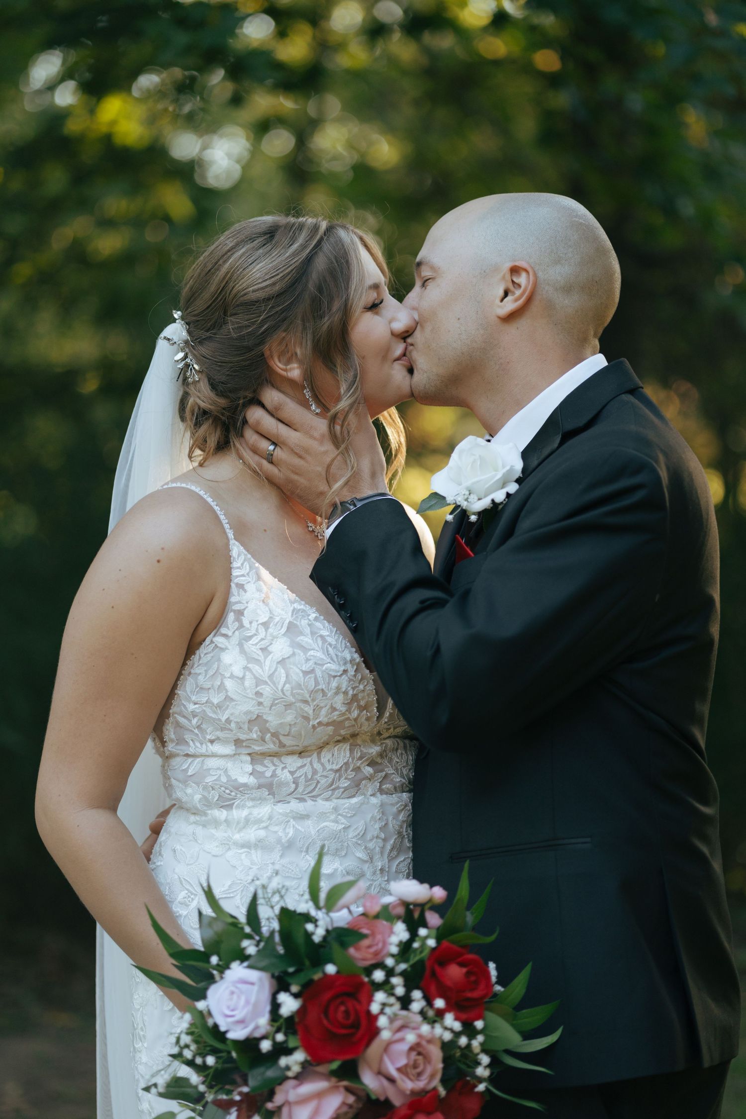 Groom tenderly kisses his bride while she holds a bouquet of red and pink roses outdoors.