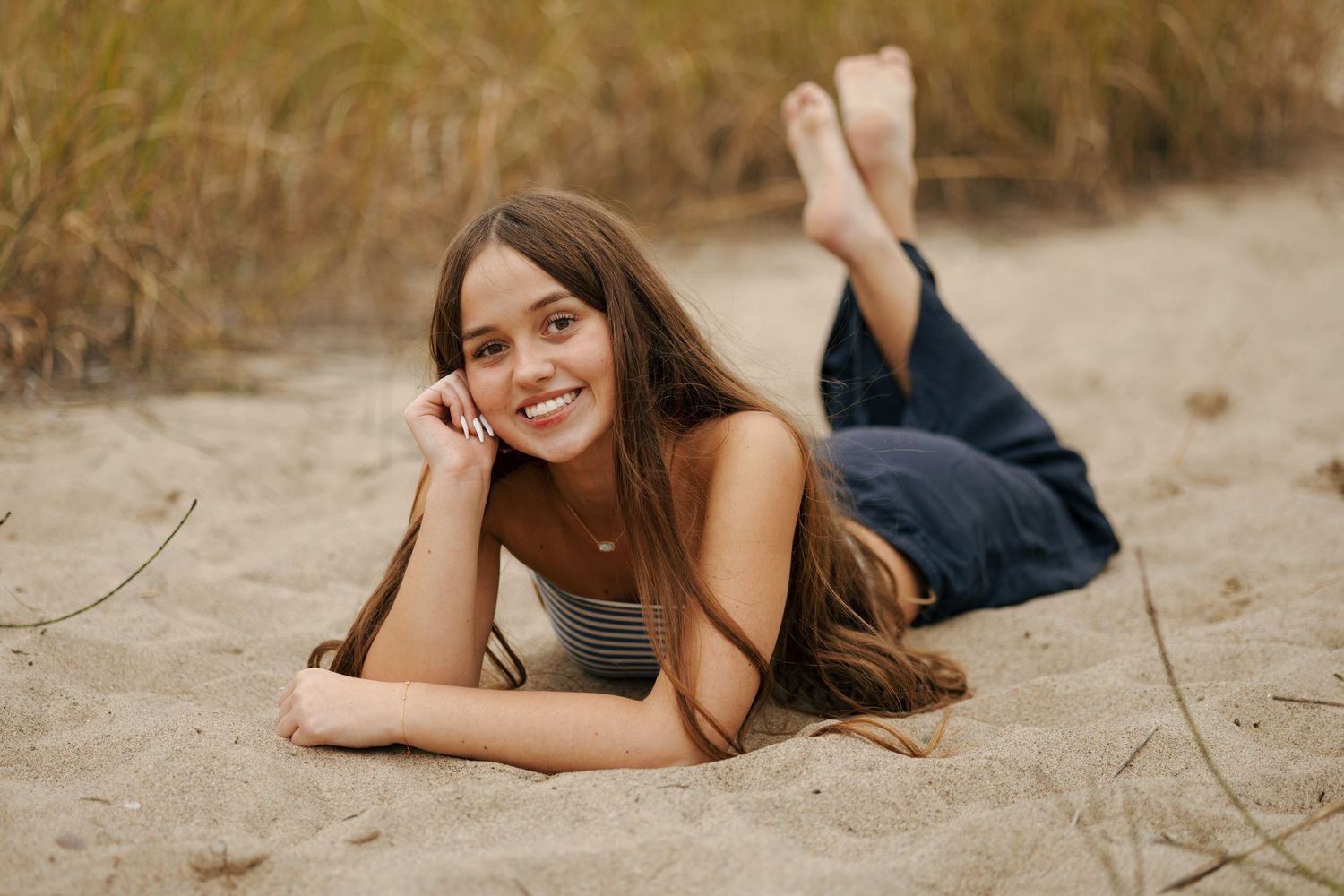 Young woman with long brown hair lying on sandy beach, smiling, wearing striped top and jeans.