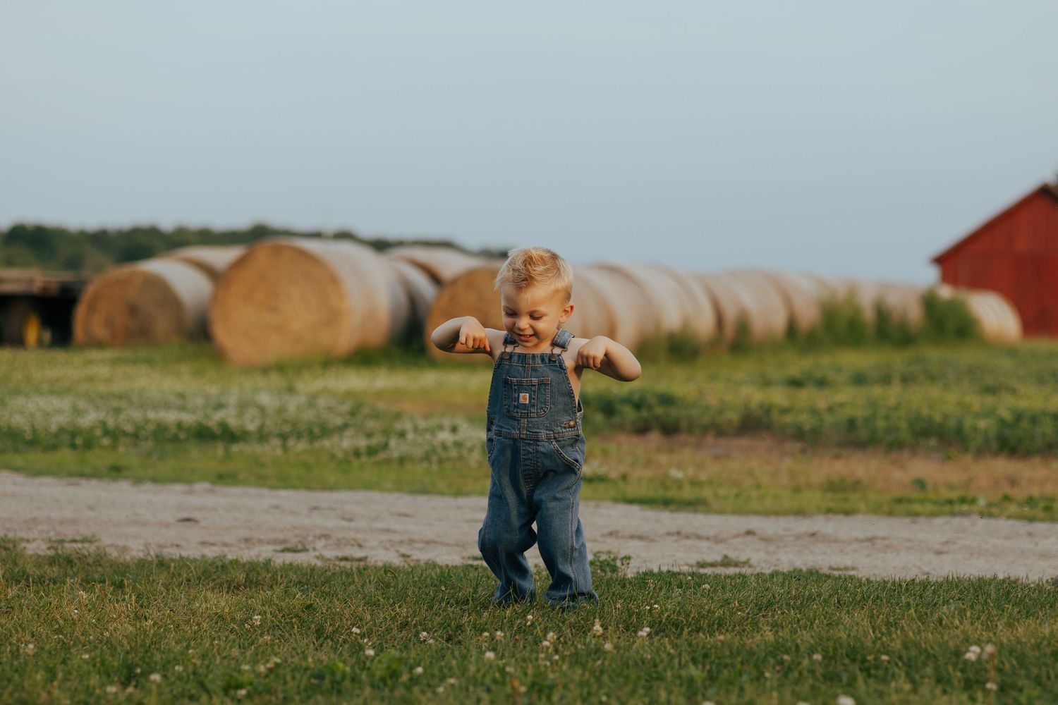 Toddler in denim overalls walking on a farm path with hay bales and red barn in background.