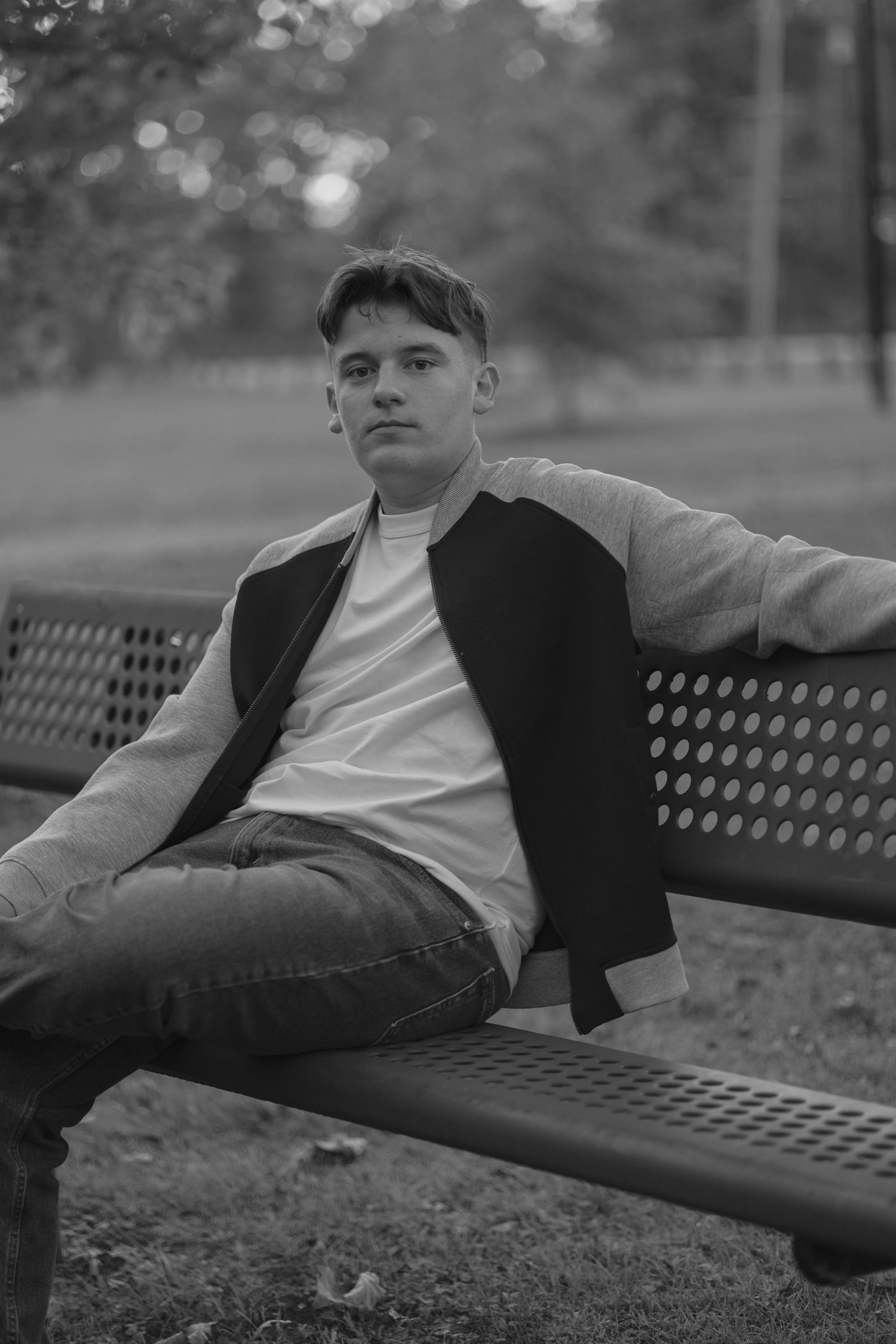 Young man in varsity jacket sitting relaxed on metal park bench in black and white portrait photo.