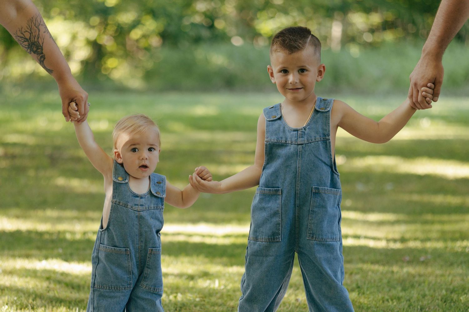Two brothers in matching denim overalls holding hands with adults in sunny green park.