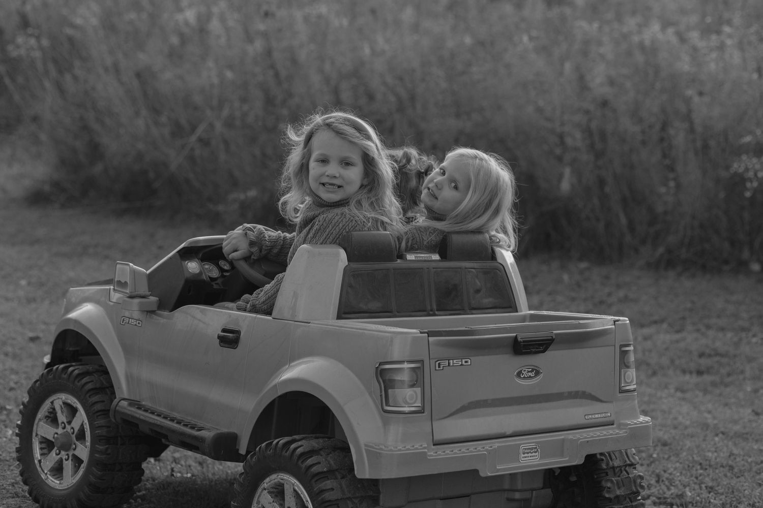 Two young girls smiling and playing in a toy Ford pickup truck outdoors in a grassy field.