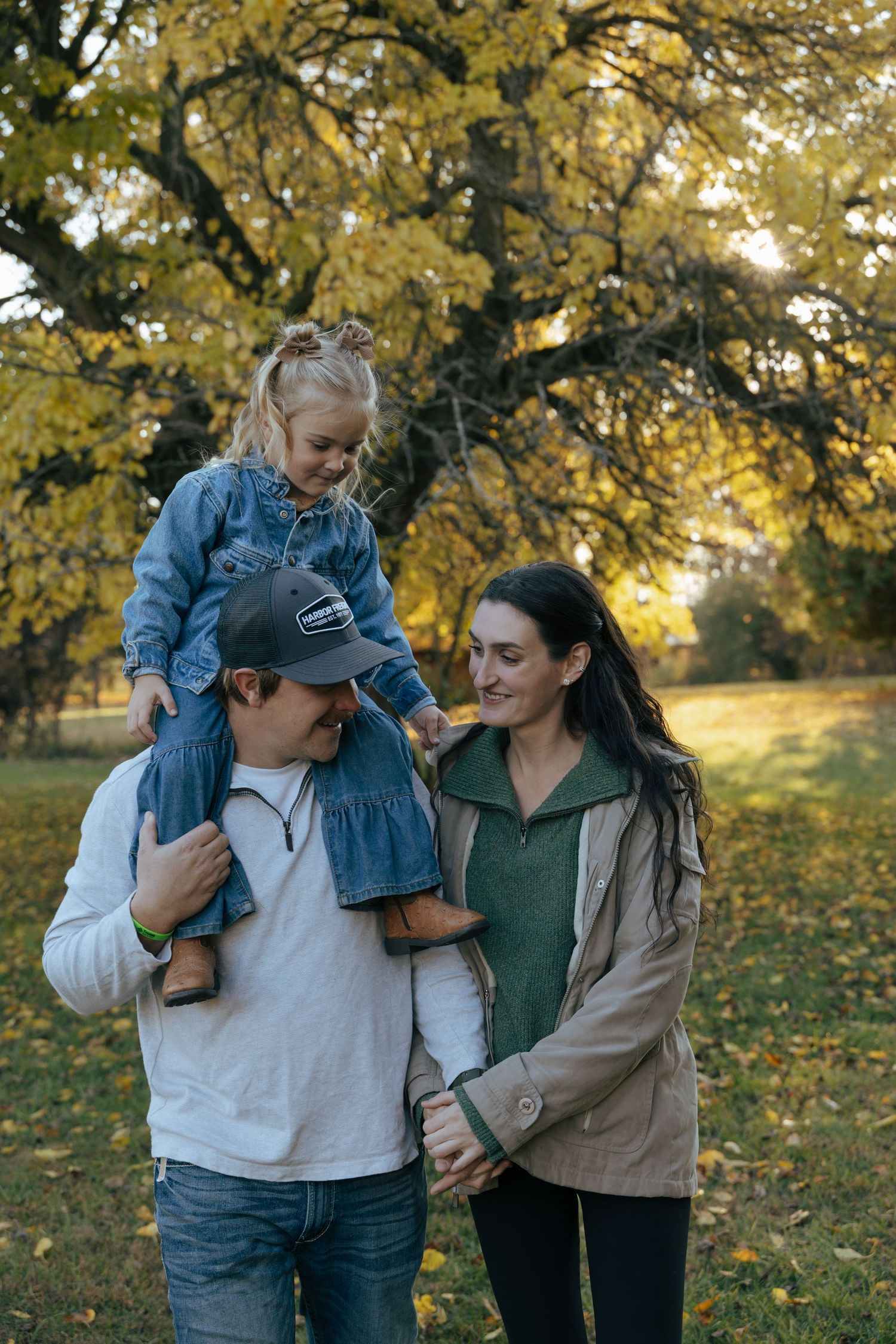Happy family with young daughter sitting on dad's shoulders walking in autumn park with golden trees.