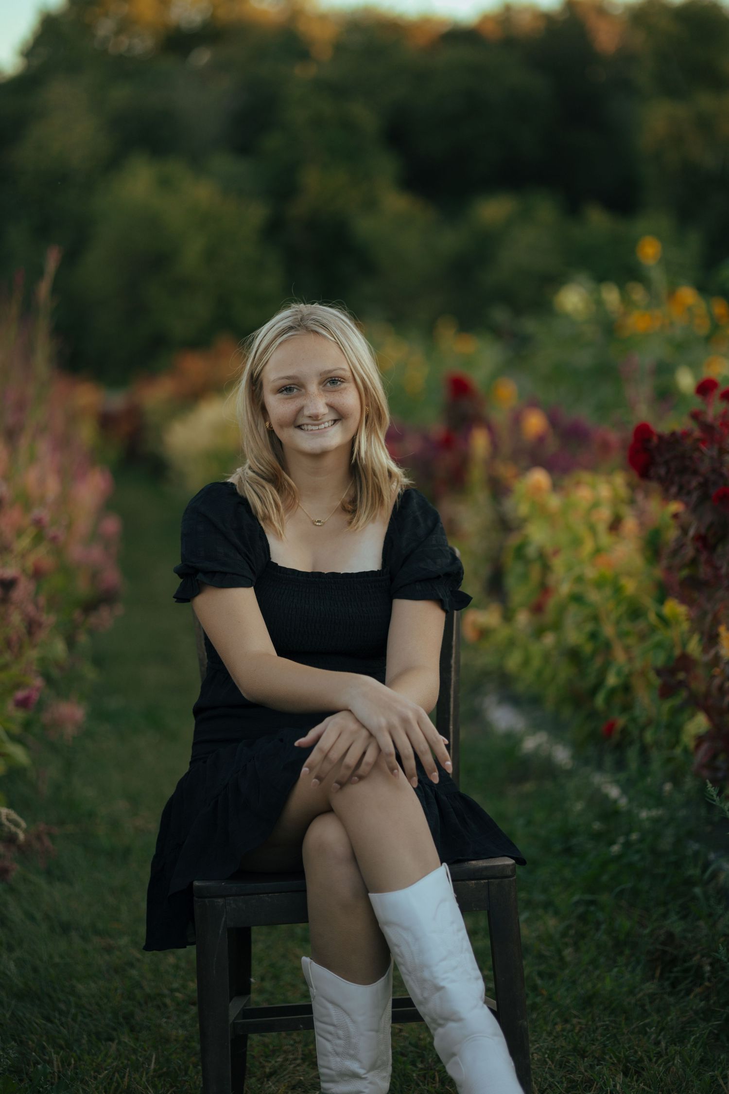 Smiling blonde teen girl in black dress and white boots sitting on chair in flower garden.