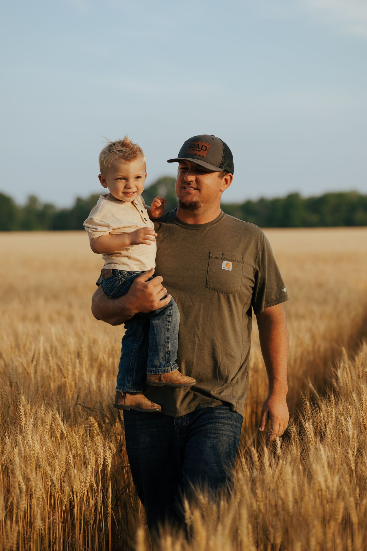 Father carrying smiling toddler through tall golden wheat field on a sunny day.