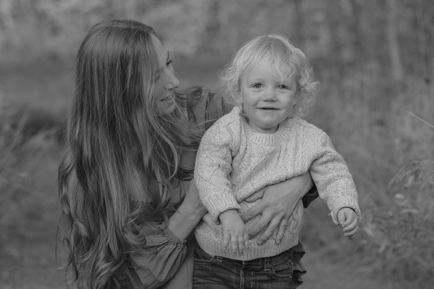 Black and white portrait of mother holding smiling blonde toddler outdoors in natural setting.
