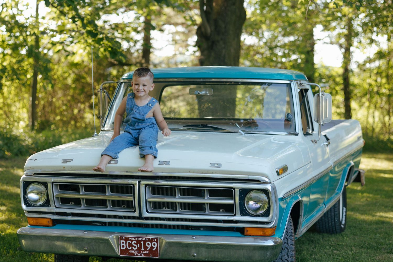 Smiling toddler in denim overalls sitting on hood of classic teal and white Ford truck.