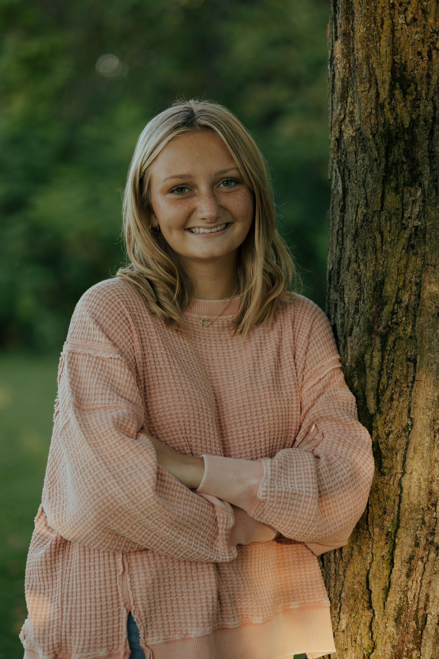 Smiling blonde teen girl in pink waffle knit sweater leaning against tree trunk outdoors.