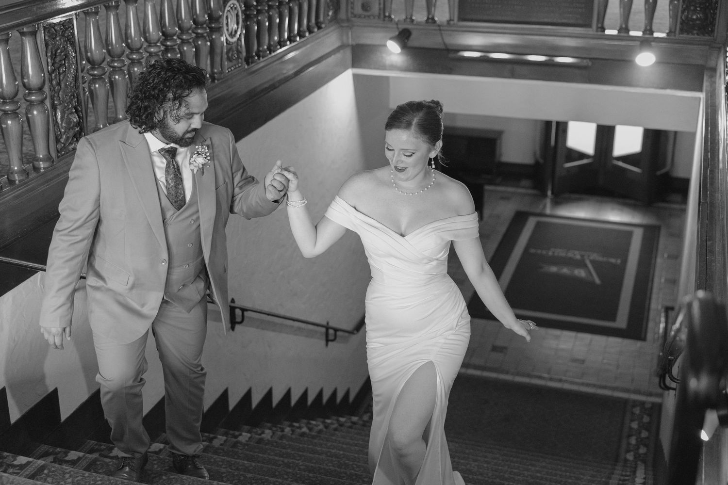 Bride and groom holding hands walking down elegant staircase in black and white wedding photo.