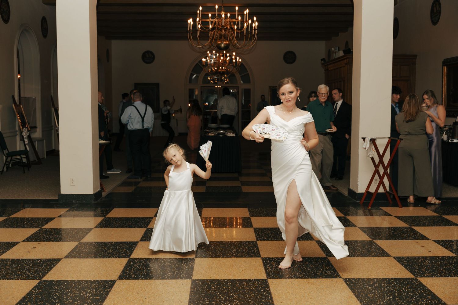 Bride in white gown dancing with young flower girl on checkered ballroom floor.