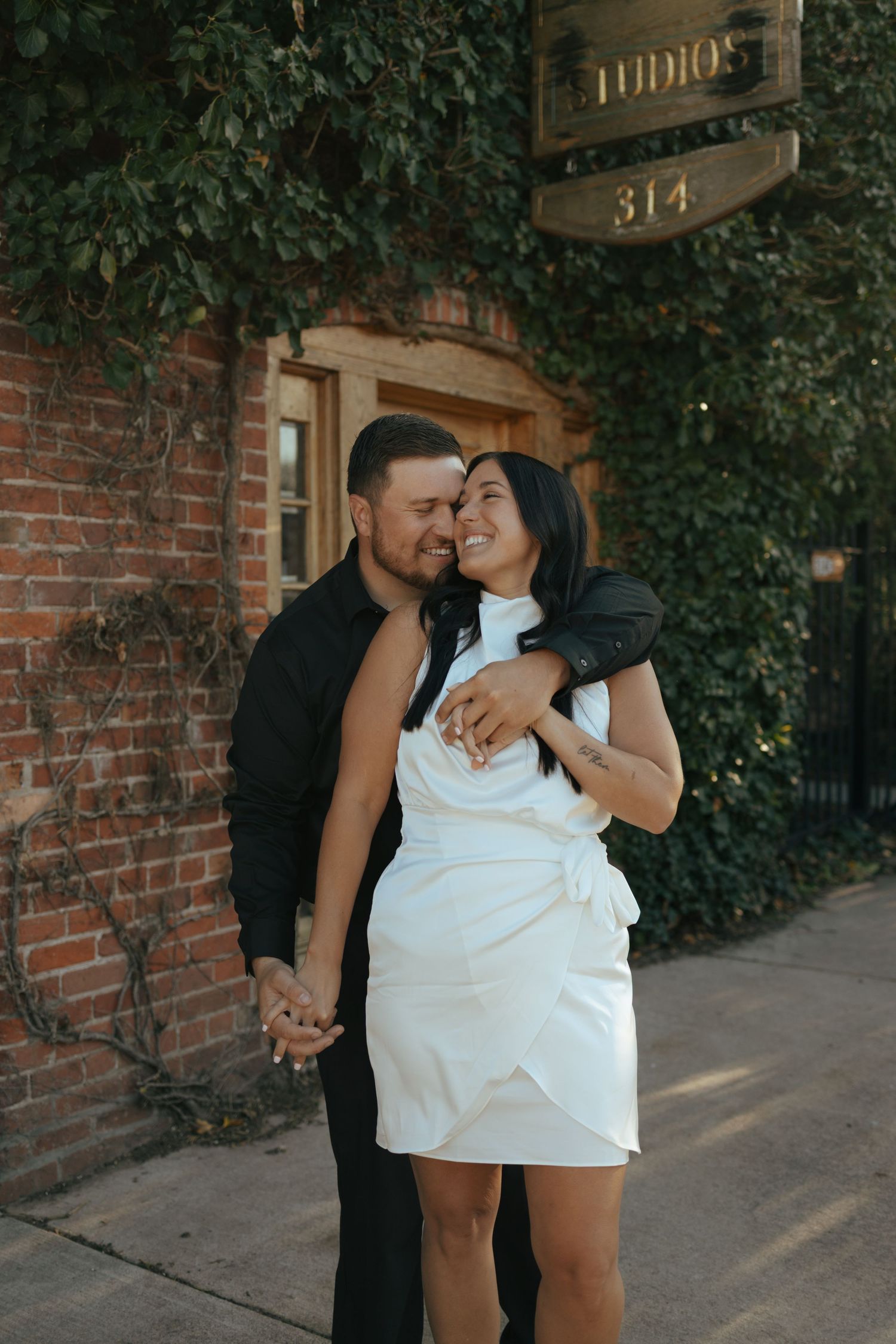 Couple embracing and smiling outside brick building covered in ivy at dusk.