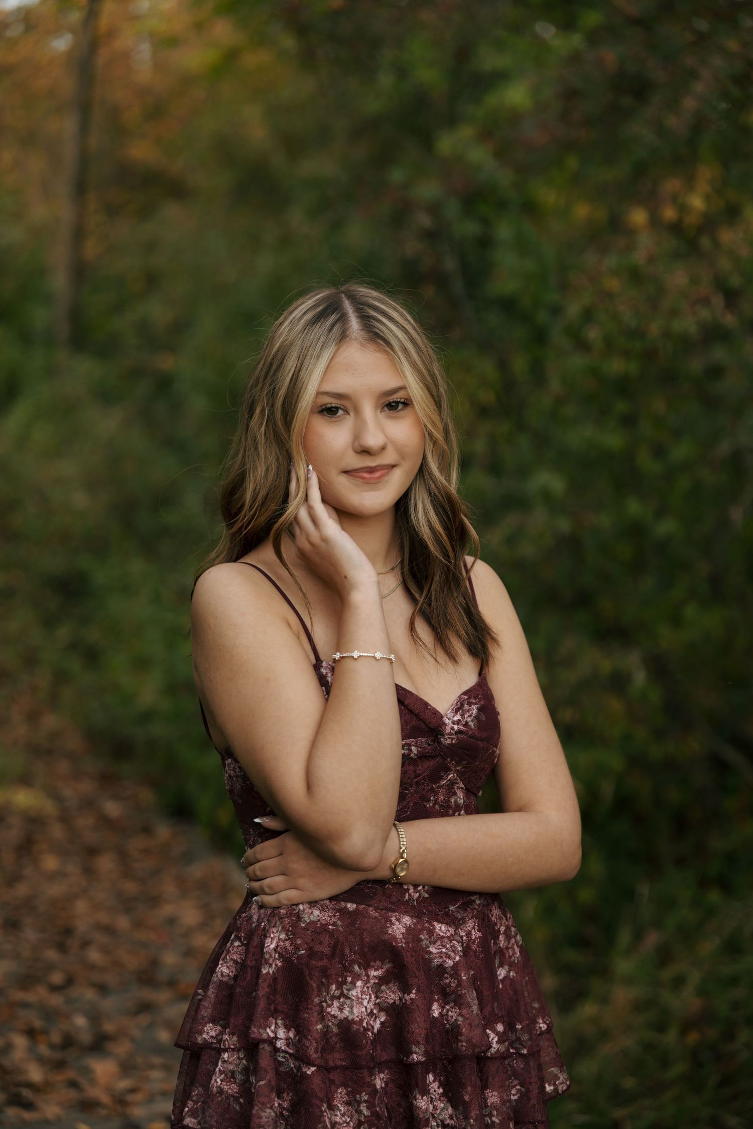 Teen girl in burgundy floral dress posing on autumn forest path with hand near face and warm fall colors behind her.