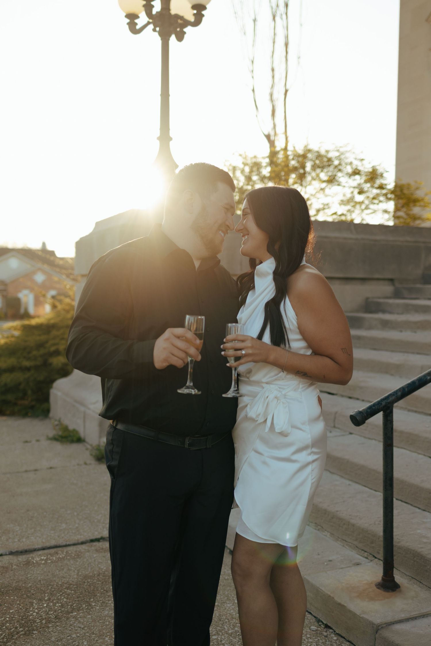 Couple sharing a romantic kiss while holding champagne glasses at golden hour on stone steps.