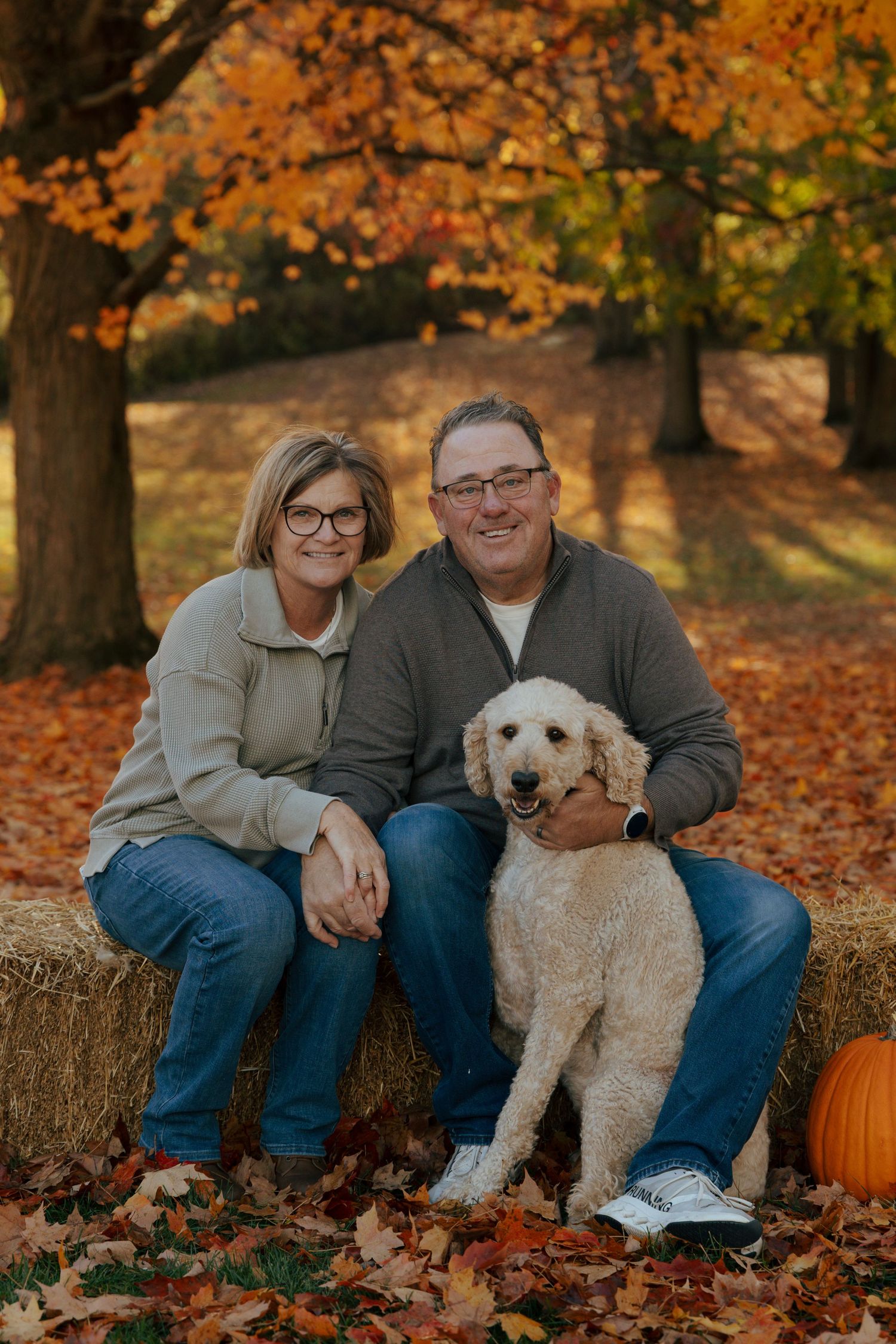 Smiling couple sitting on hay bale with fluffy golden doodle dog amid fall pumpkins and leaves.