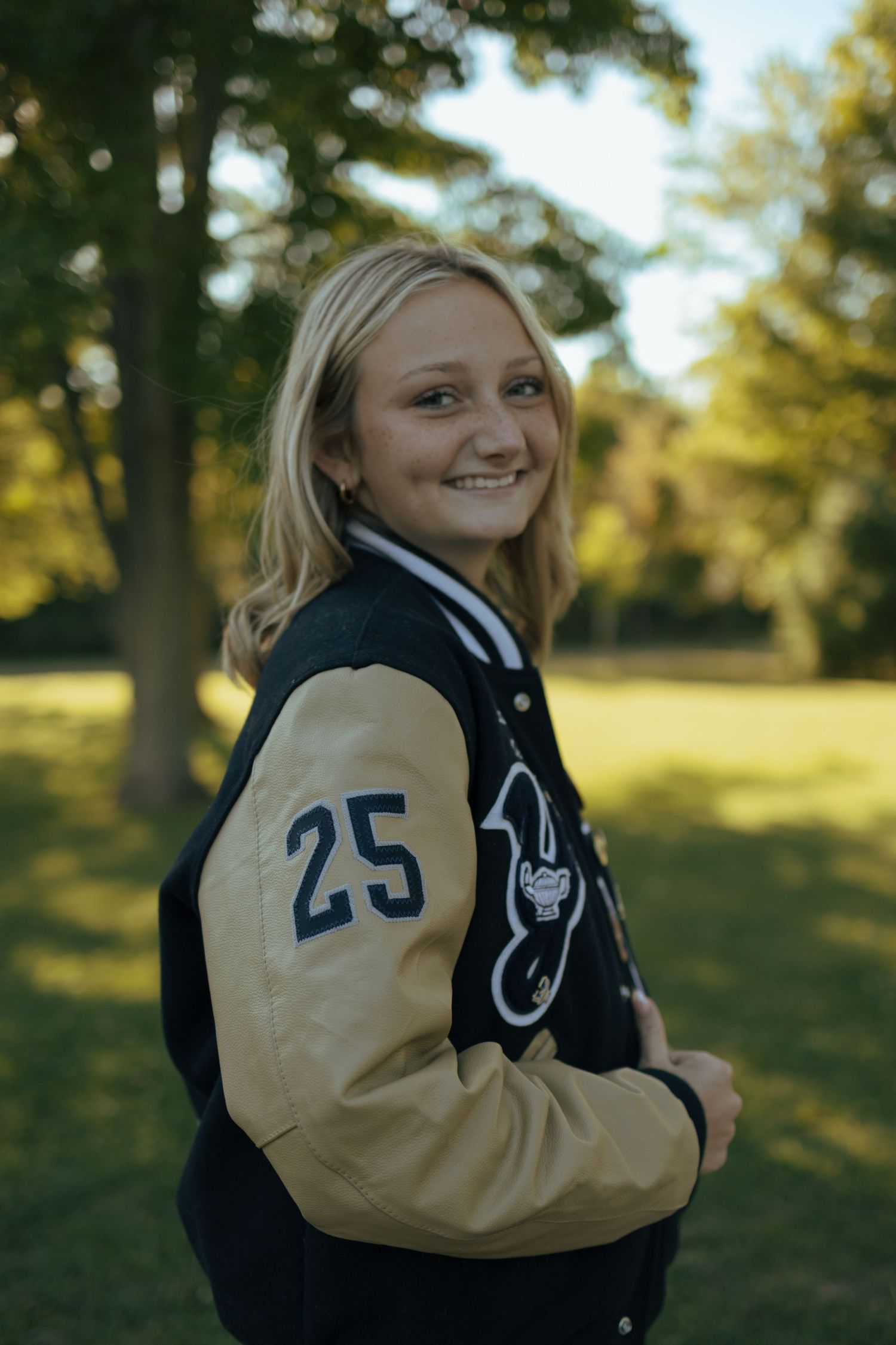 Smiling blonde girl in navy and gold varsity jacket with number 25 posing outdoors in green park.