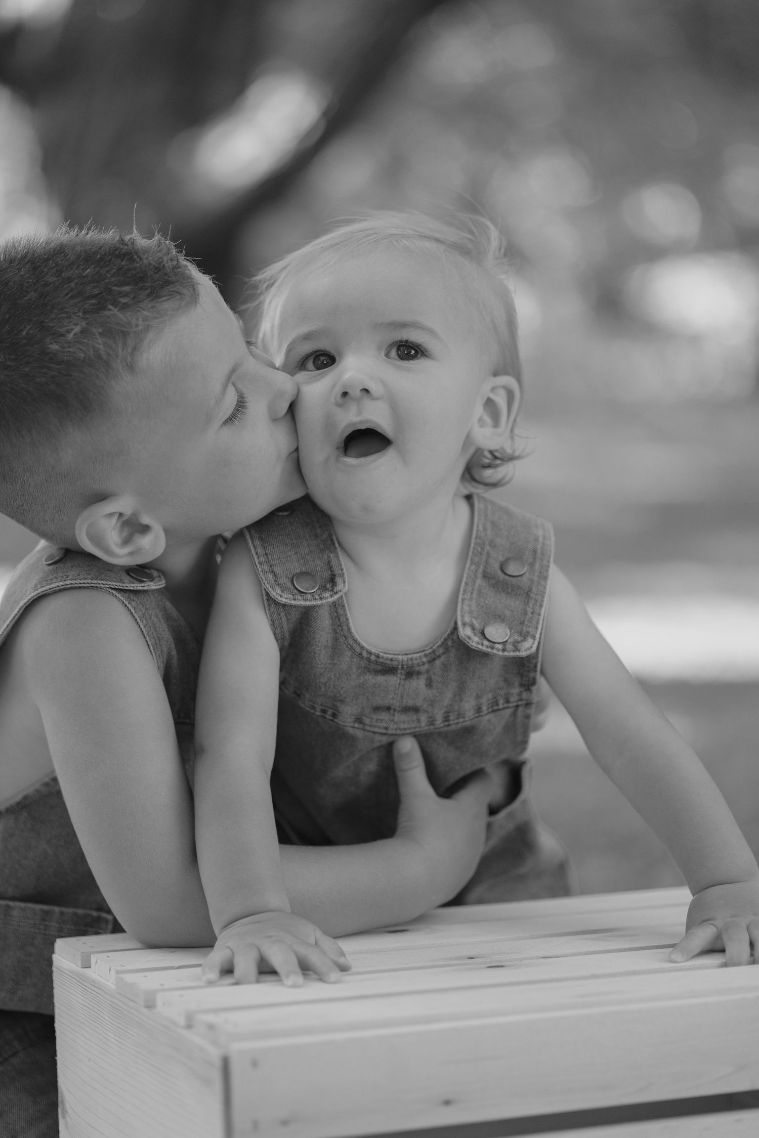 Older boy kissing surprised baby sibling on cheek, black and white photo.