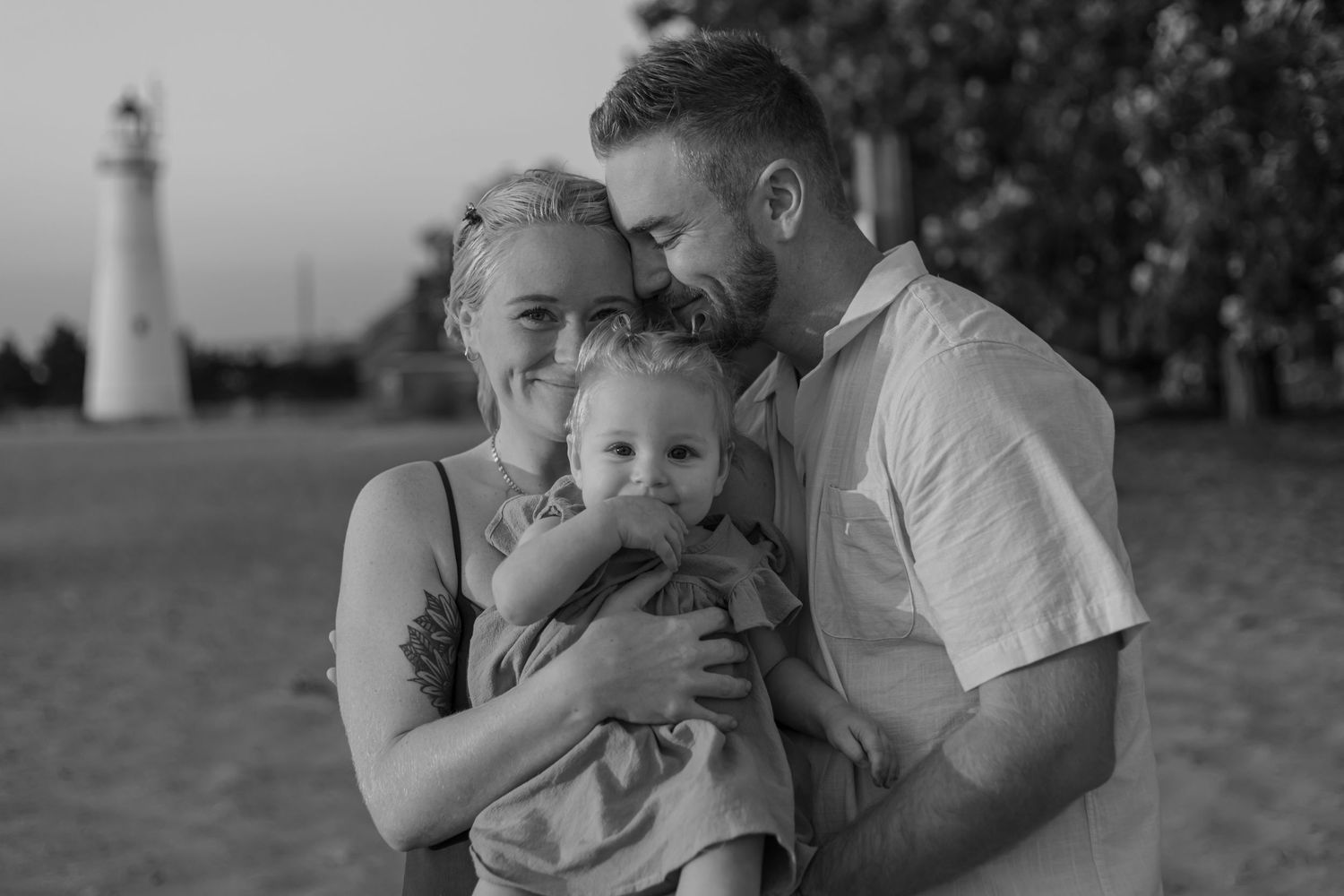 A black and white portrait of a loving couple with their baby near a lighthouse at the beach.