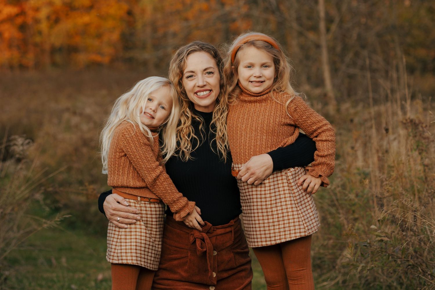 Smiling mother posing with two young blonde girls in coordinating brown plaid outfits in autumn park.