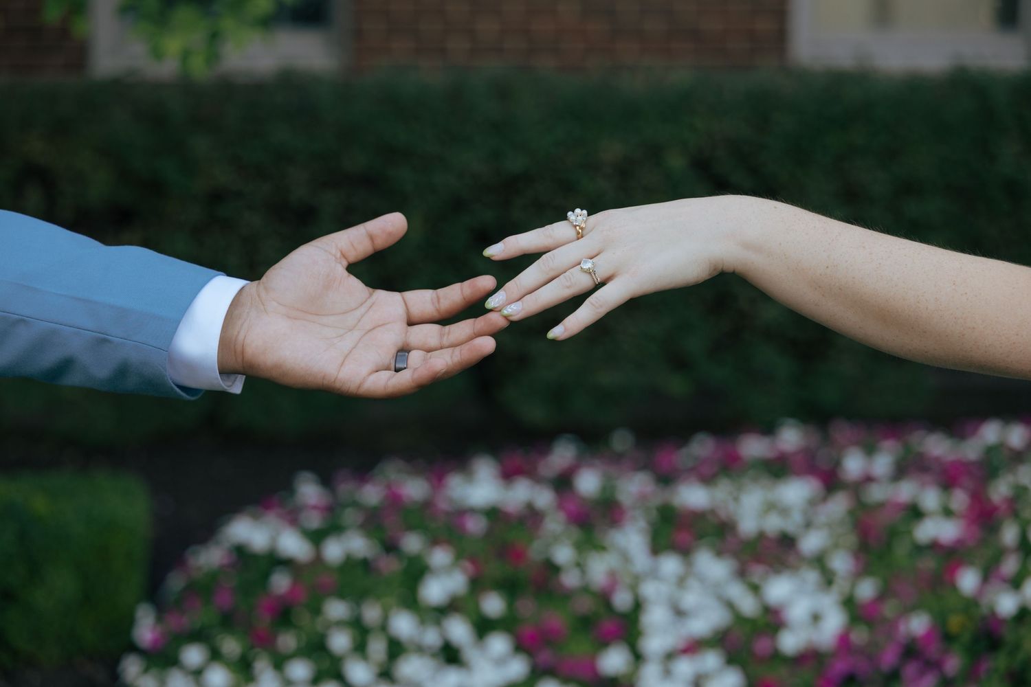 Close-up of bride and groom reaching hands toward each other, showing wedding rings near flower garden.