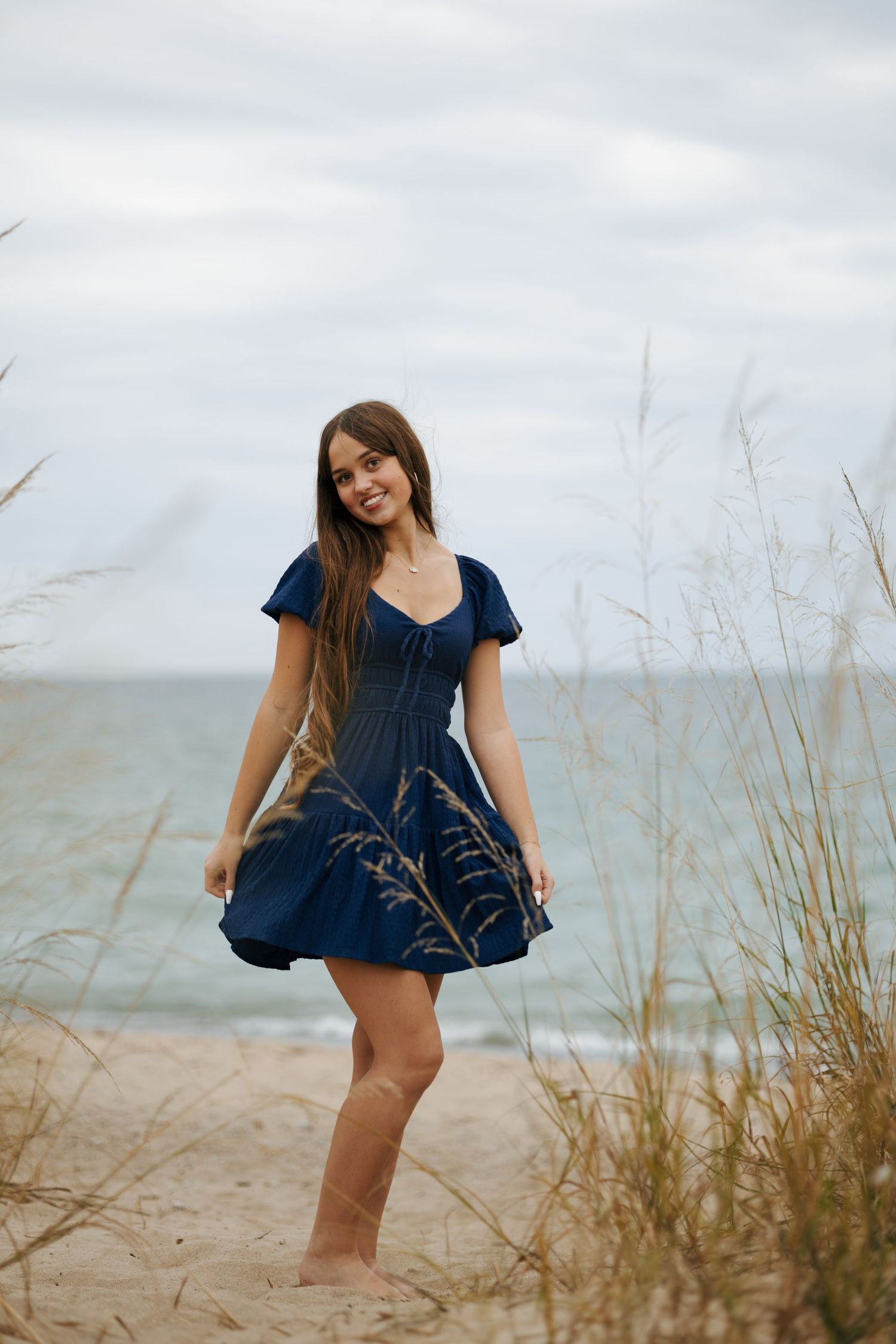 Smiling young woman in navy blue dress standing on sandy beach among tall grass with ocean backdrop.