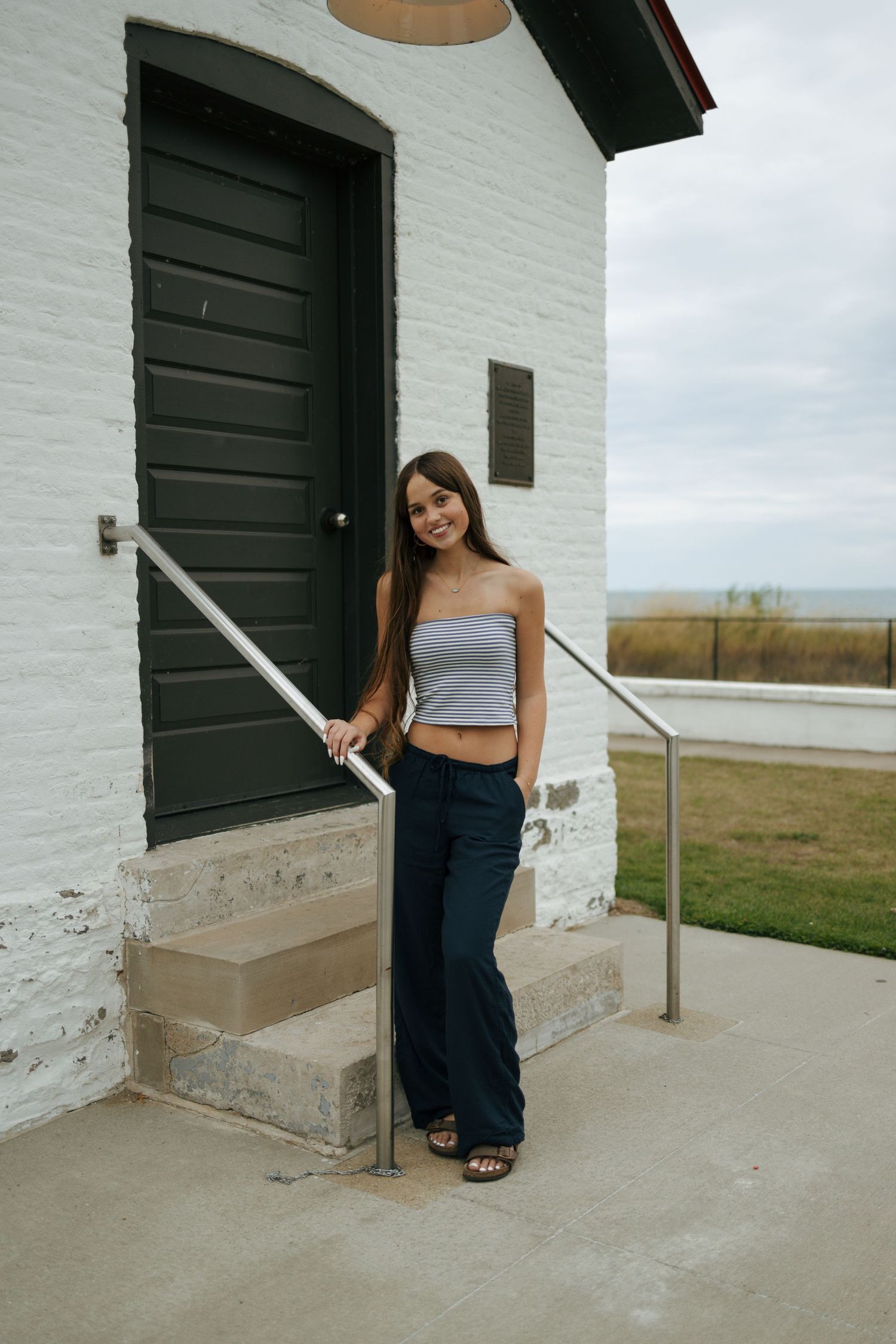 Smiling young woman in gray tube top and wide-leg pants posing on steps of white brick building.