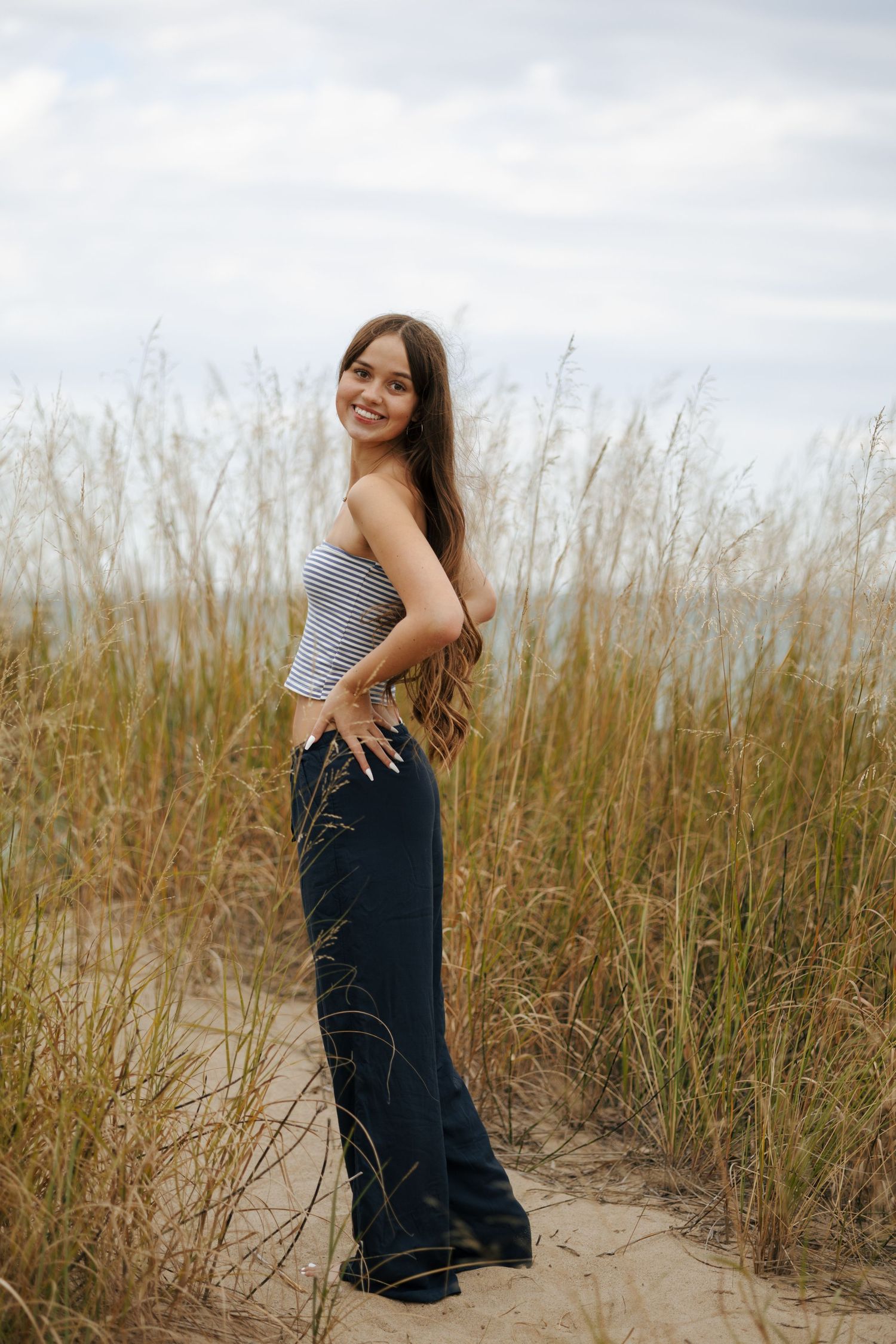 Young woman in striped crop top and dark pants standing among tall beach grass, smiling over shoulder.