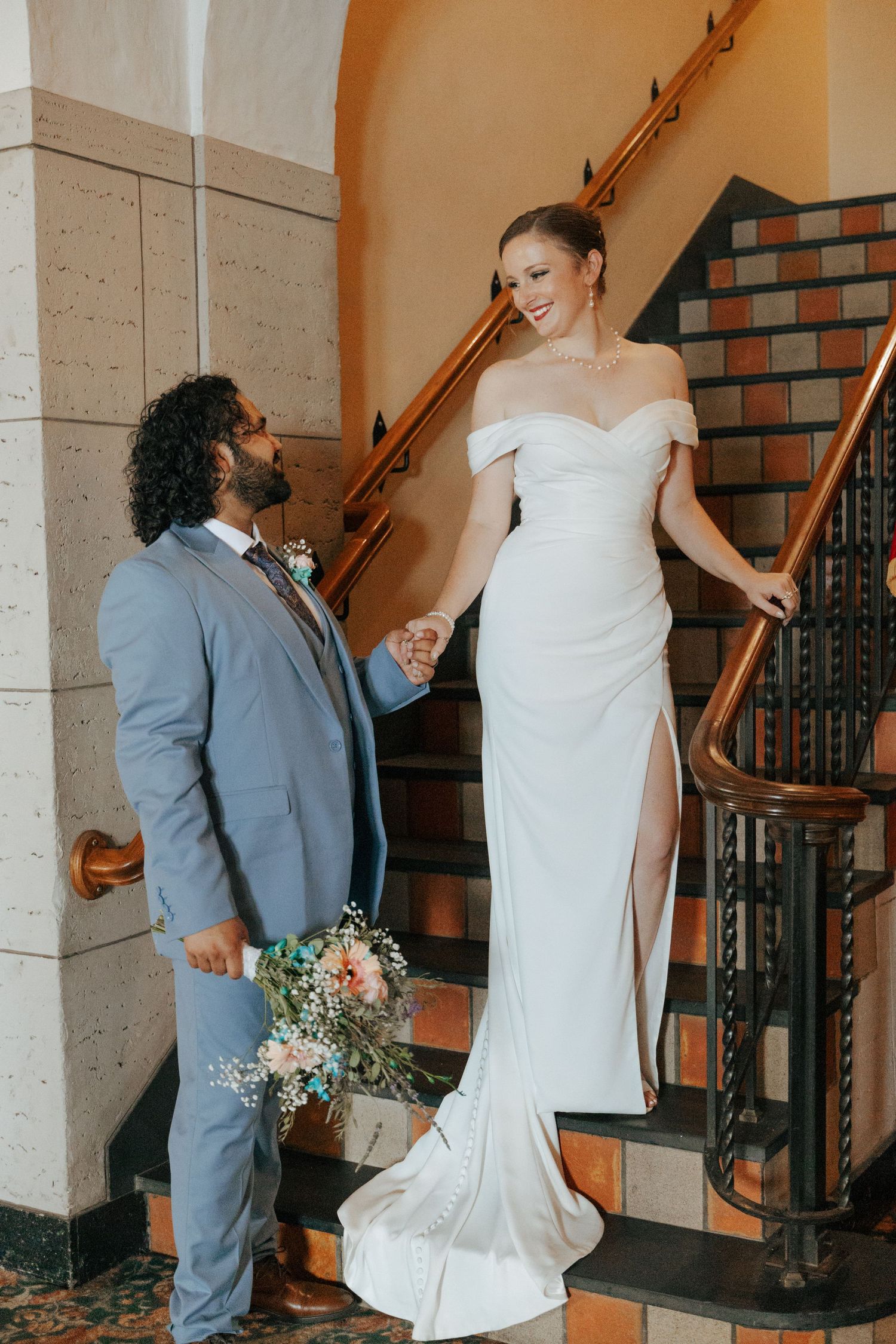 Bride and groom holding hands on ornate tiled staircase, bride in off-shoulder gown holding bouquet.
