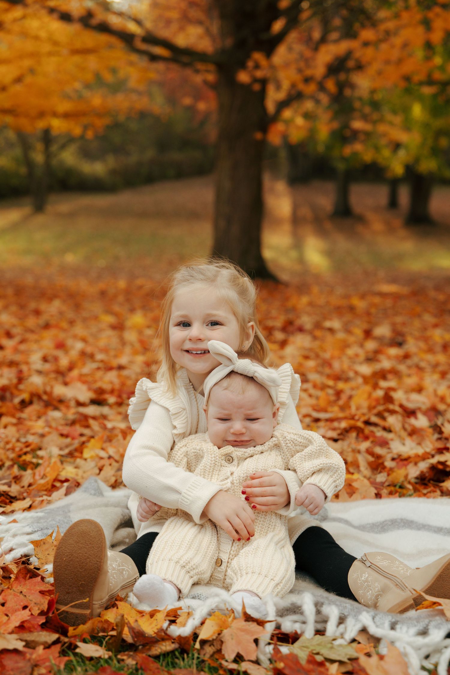 Older sister hugging baby sibling on blanket surrounded by vibrant red and orange fall leaves.
