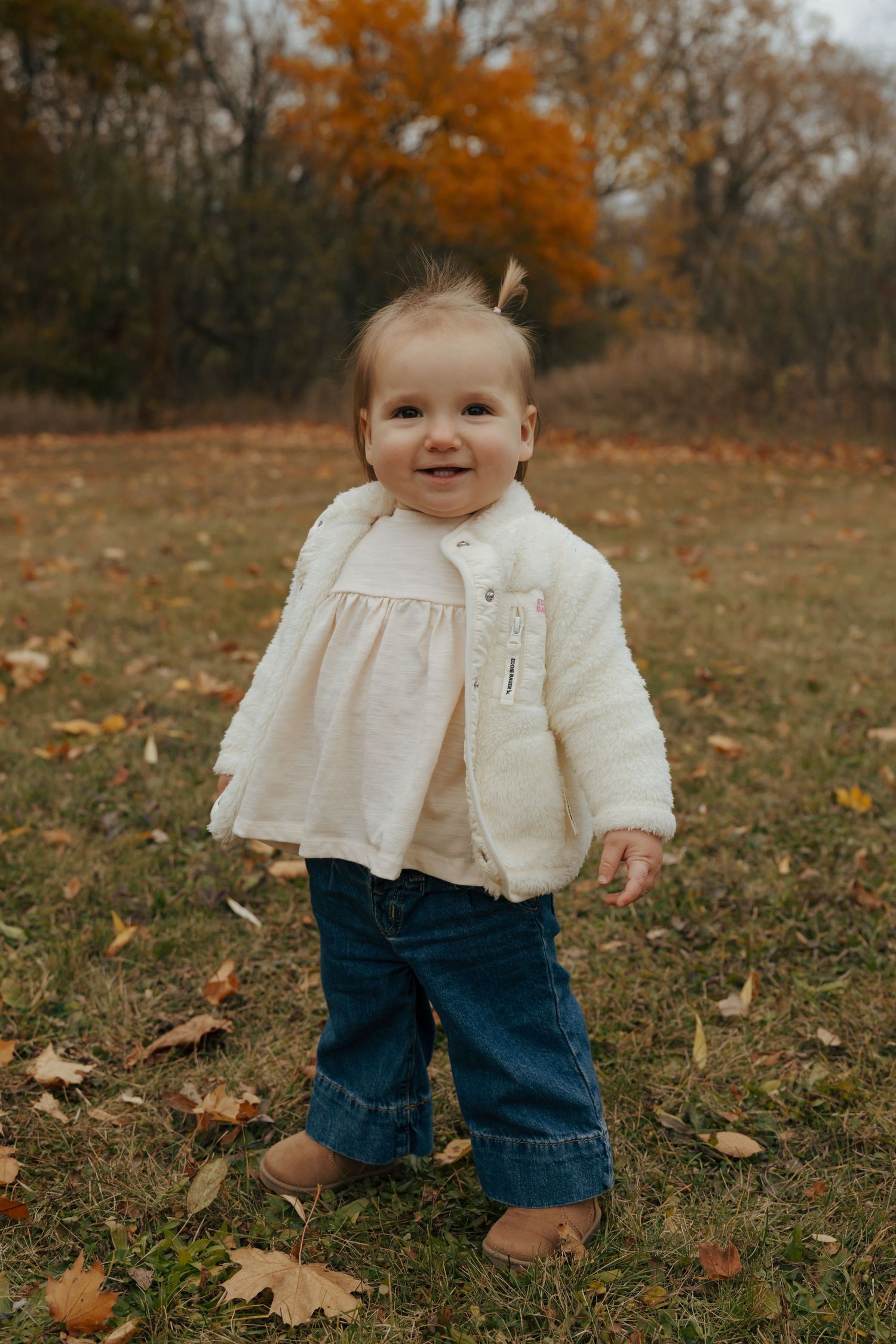 Smiling baby girl in white fuzzy jacket and jeans standing on grass with fall leaves.