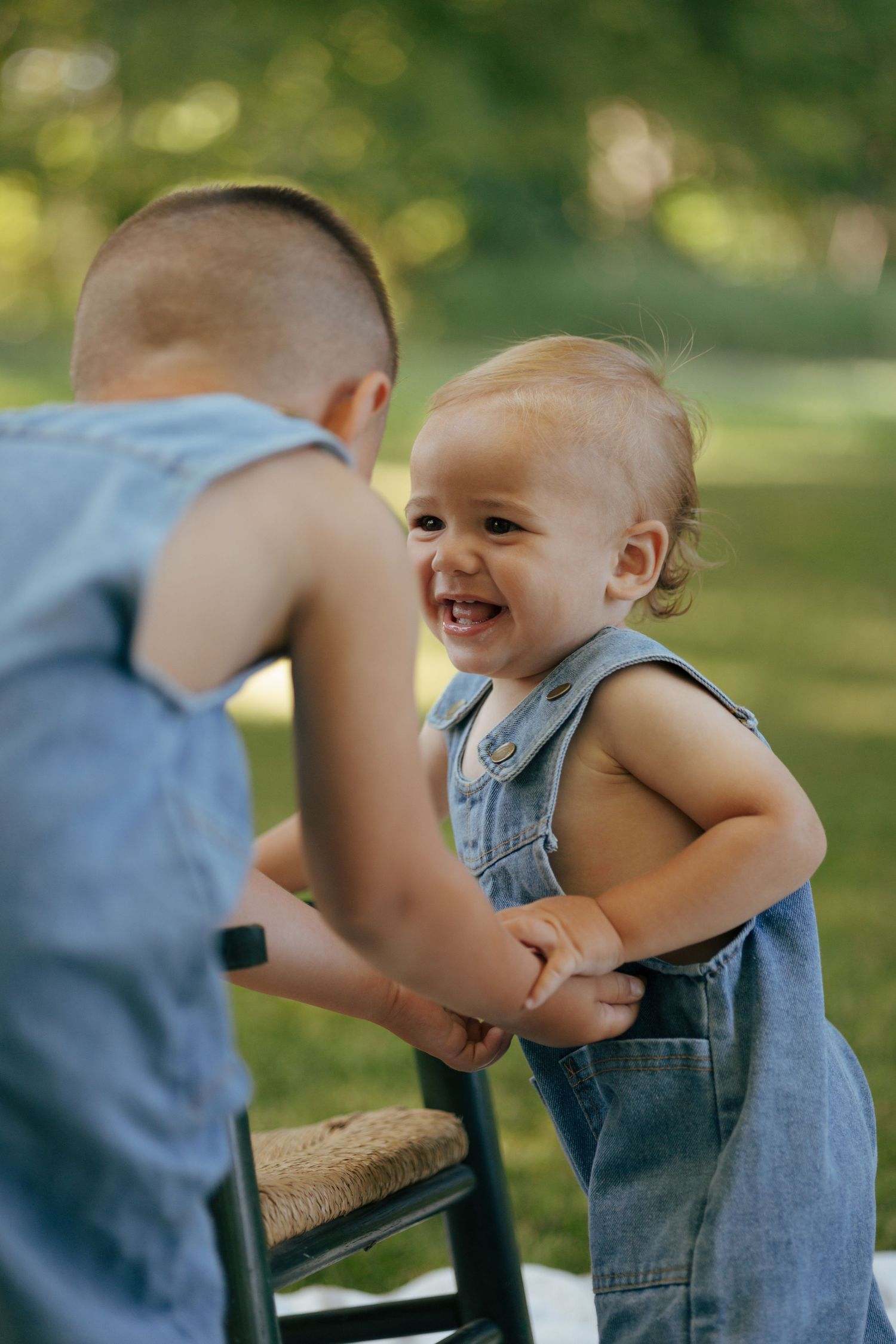 Laughing toddler in denim overalls holds hands with older sibling in a sunny green park.