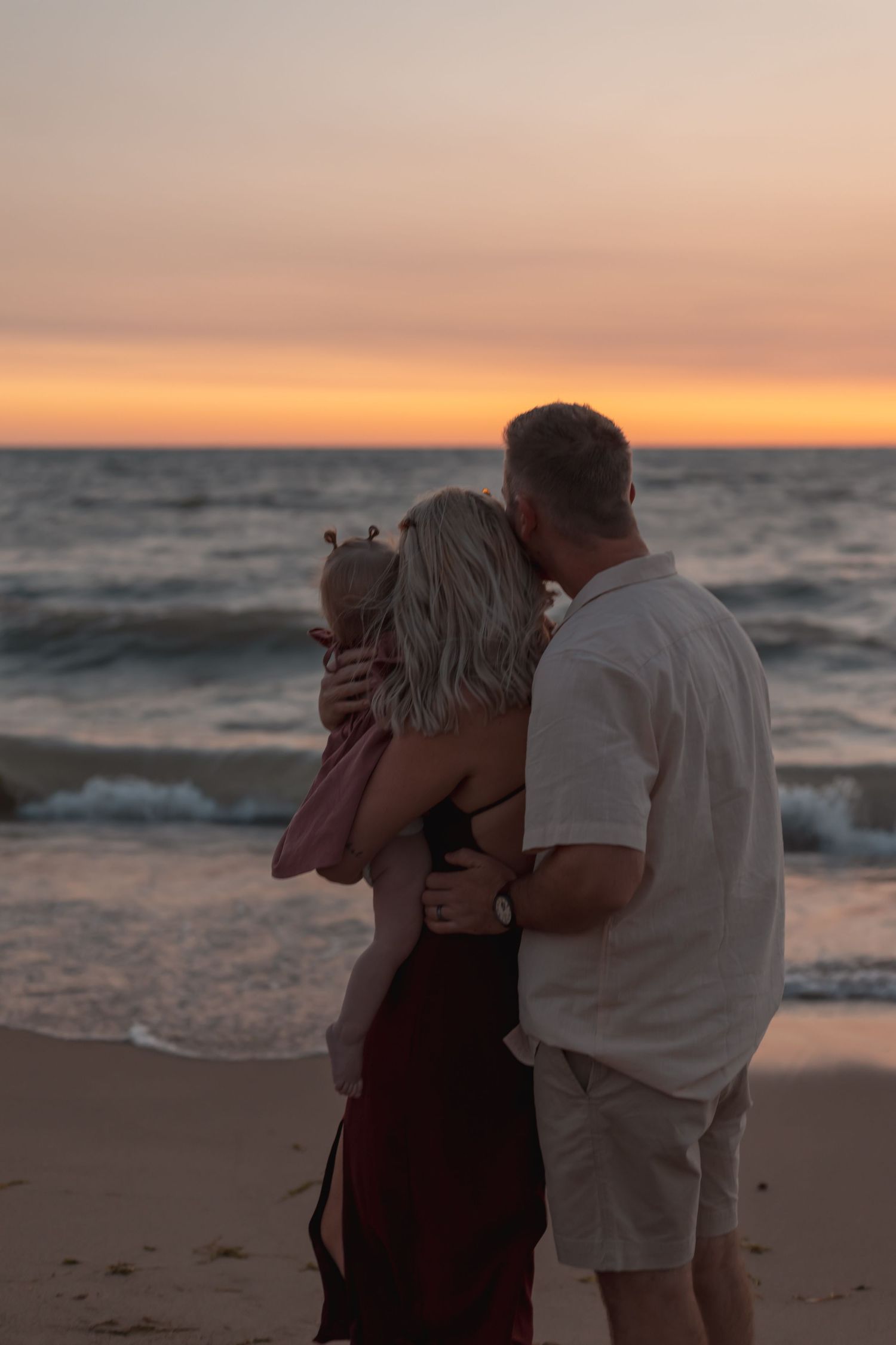 A couple embracing with their young child while watching a stunning orange sunset over the ocean.