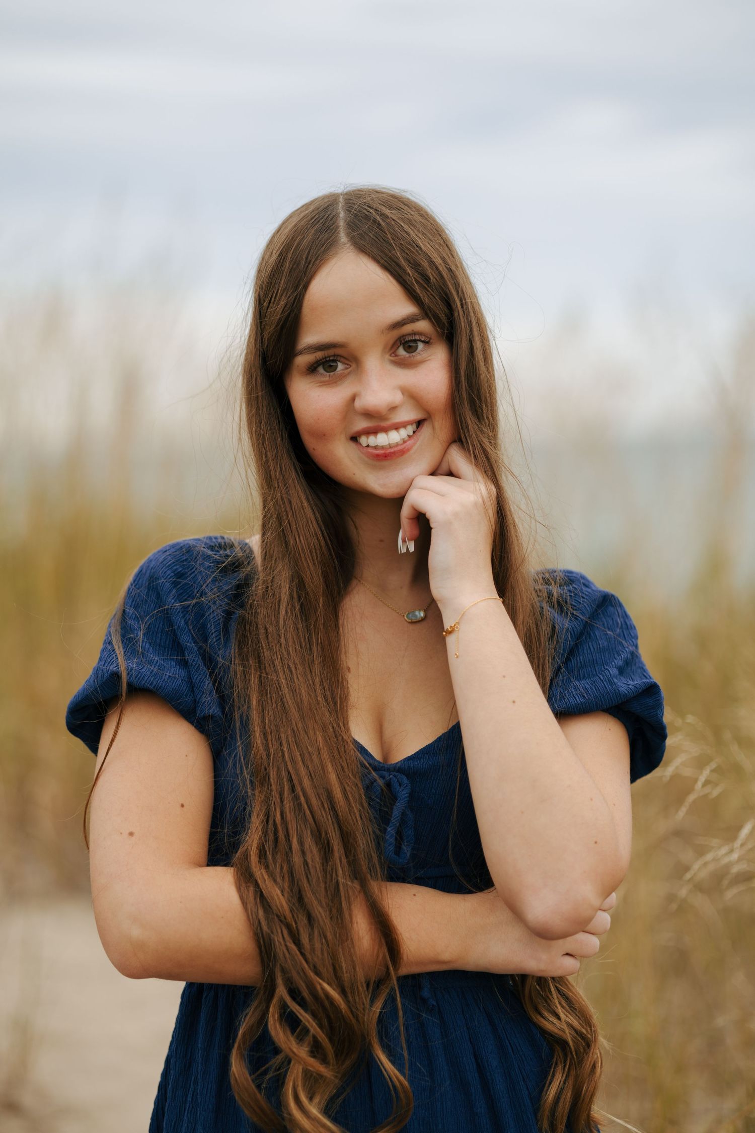 Smiling young woman with long brown hair in blue dress posing on sandy beach with tall grass.