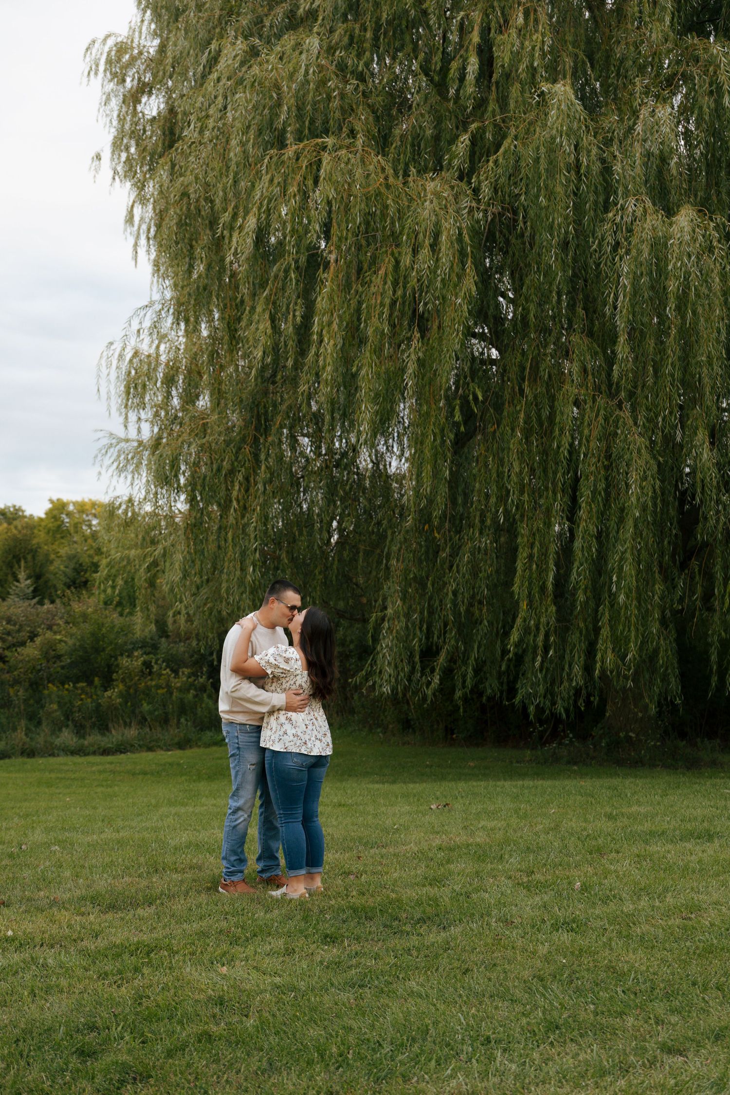 Couple sharing a romantic kiss standing in a green meadow in front of a large willow tree.