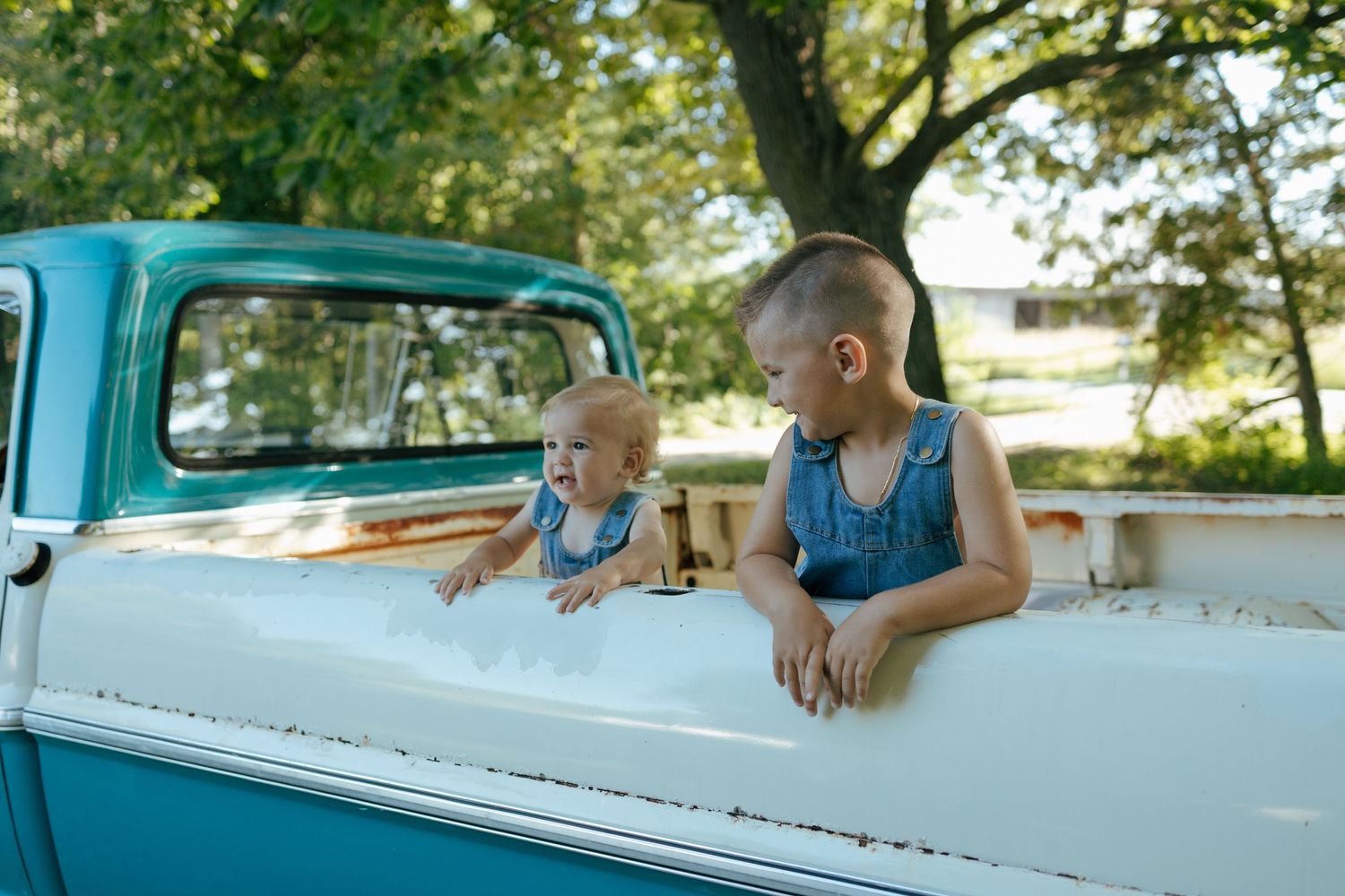 Two young children in denim overalls peek over the side of a vintage teal and white truck.