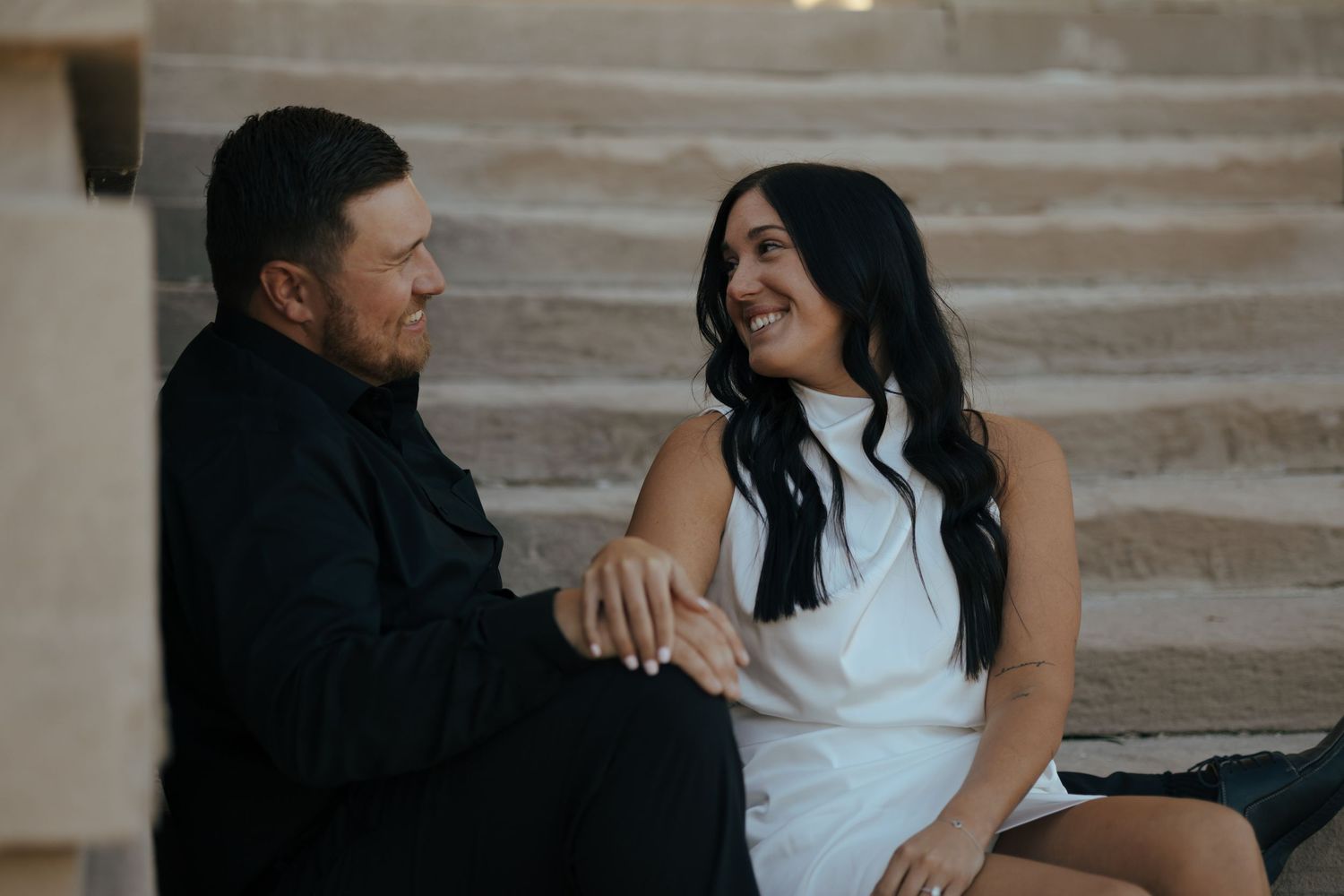 Couple sitting together on stone steps, smiling and holding hands in warm natural light.