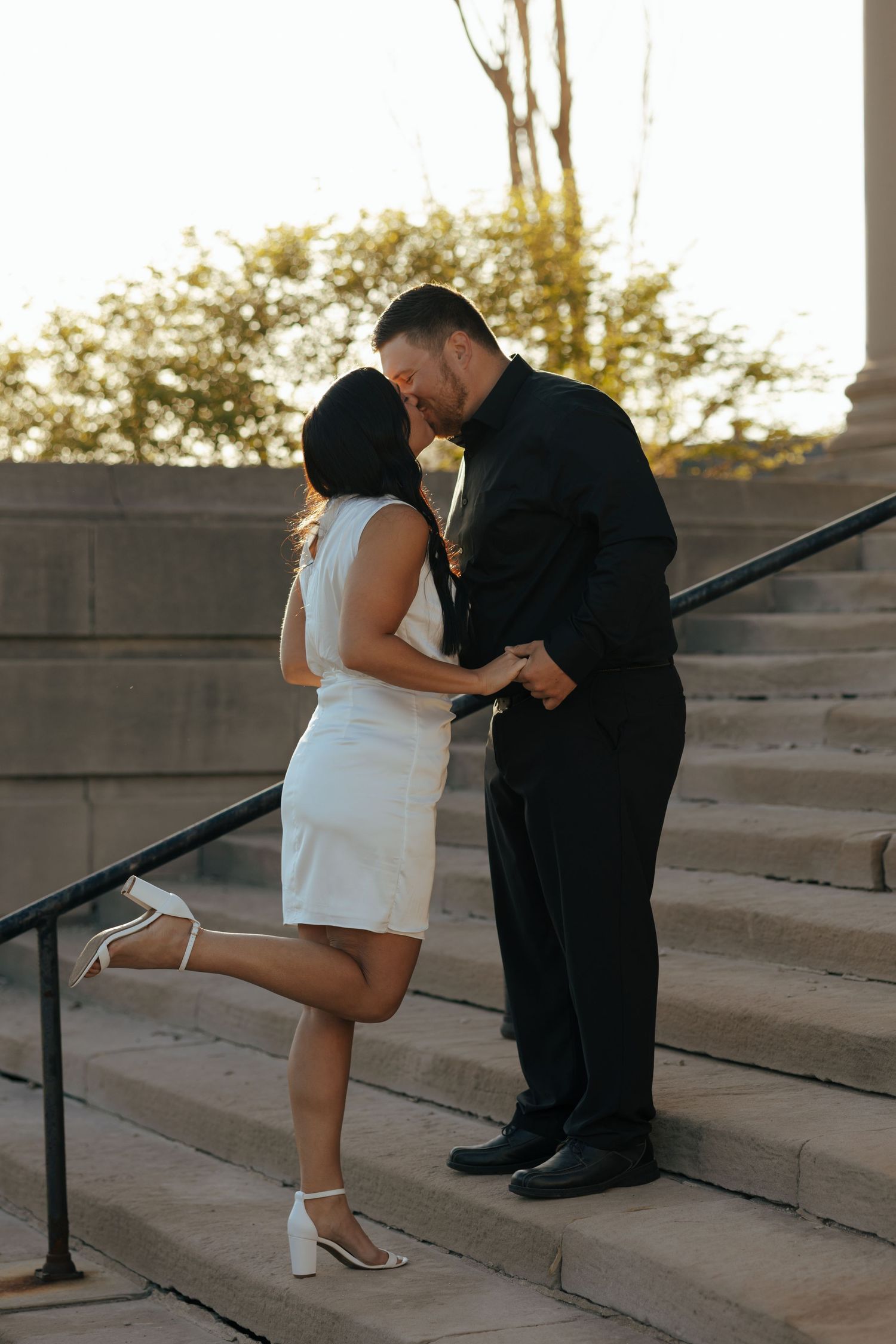 Couple sharing a kiss on stone steps at golden hour, woman lifting one leg romantically.