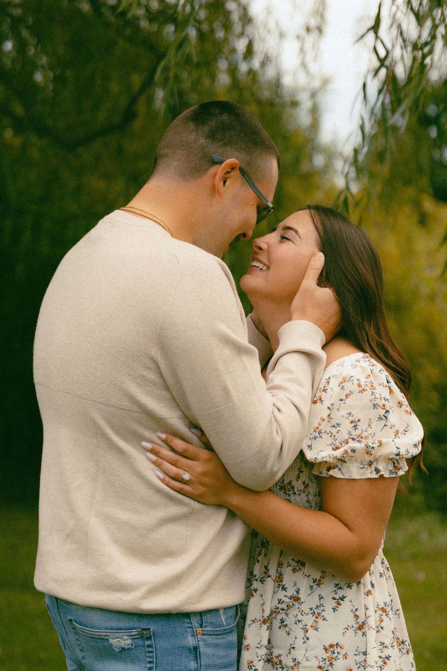 Laughing couple touching foreheads romantically beneath weeping willow tree during engagement photos.